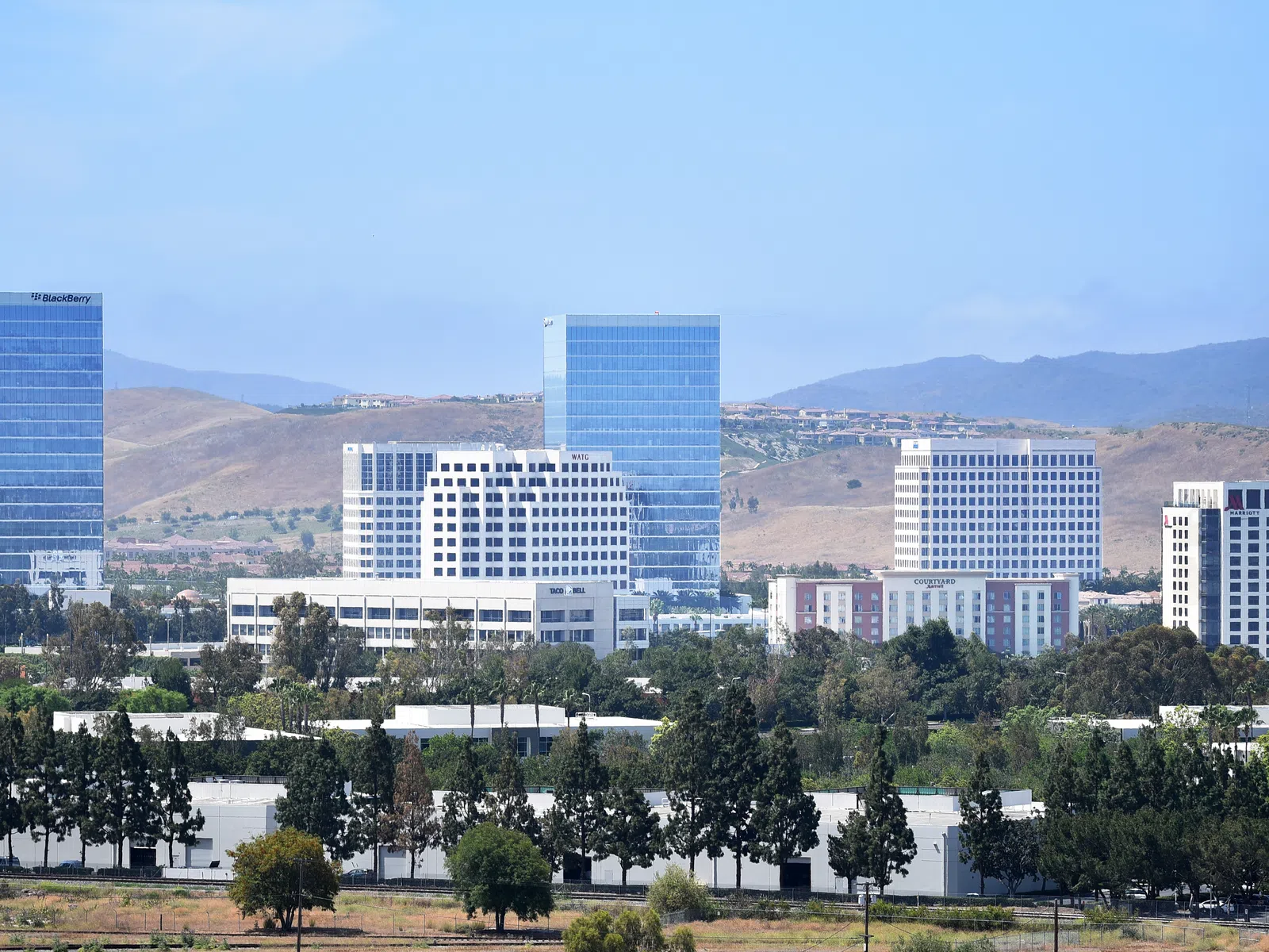 Cityscape with modern office buildings, greenery in foreground, and dry hills under clear blue sky in background