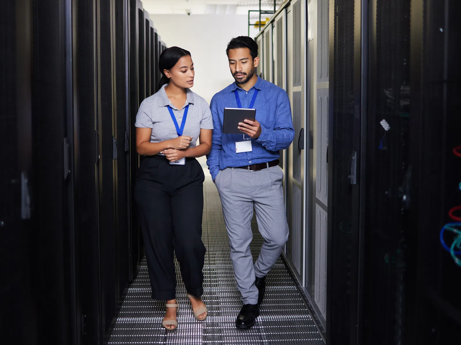 Two IT professionals walk and discuss while checking a tablet in a server room with racks on both sides.