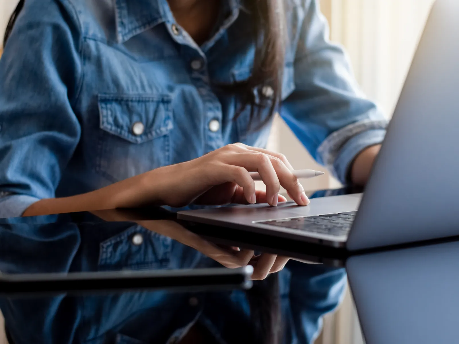 Person in denim shirt using laptop with a tablet on reflective black table in bright workspace.