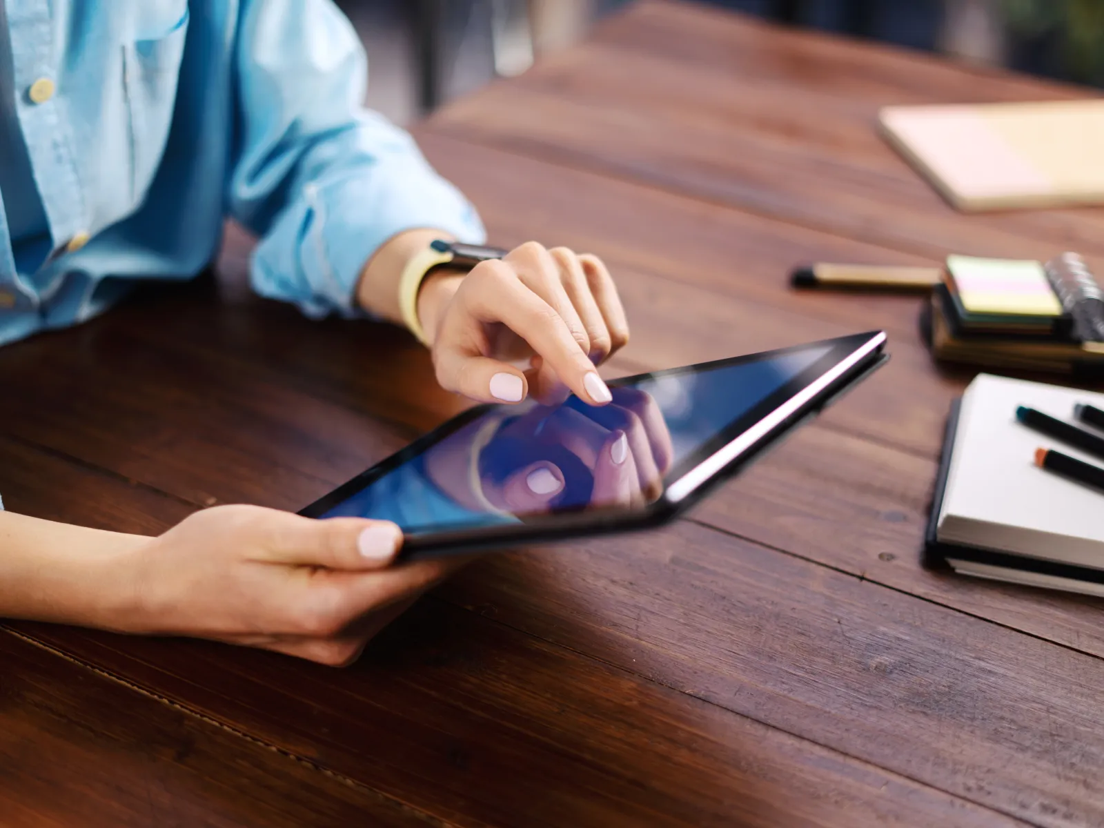 Person using a touchscreen tablet on a wooden table with notebooks and pens nearby in a casual setting