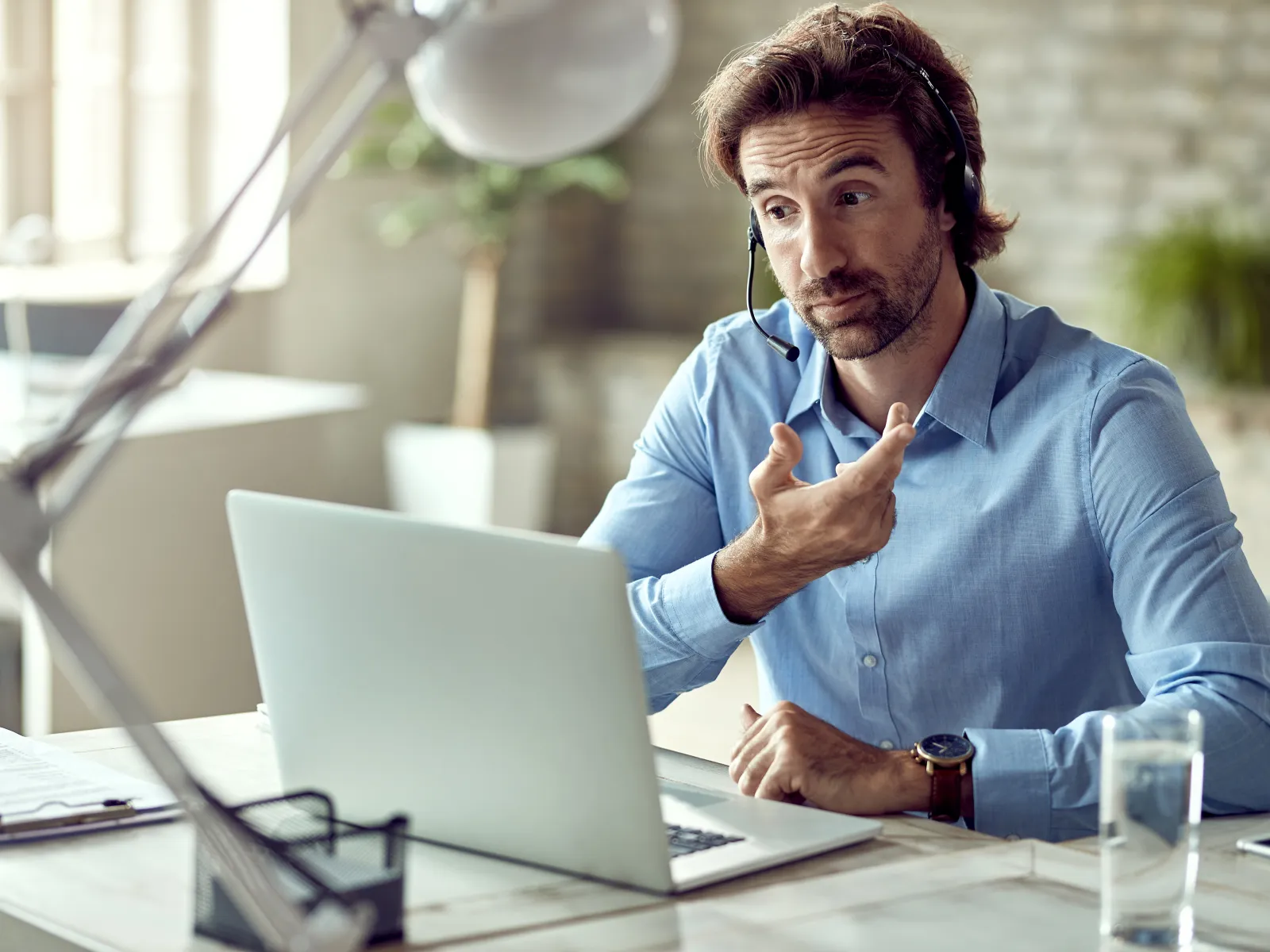 Man in blue shirt using headset for video call while working on laptop at office desk.