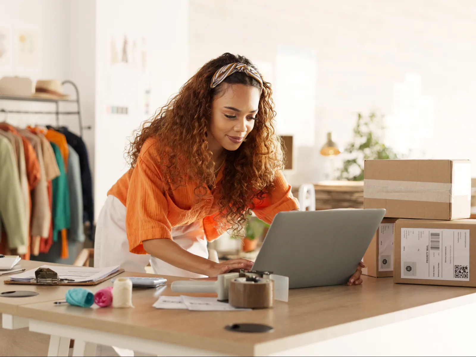 Woman in orange shirt using laptop at a workspace with clothing rack and shipping boxes