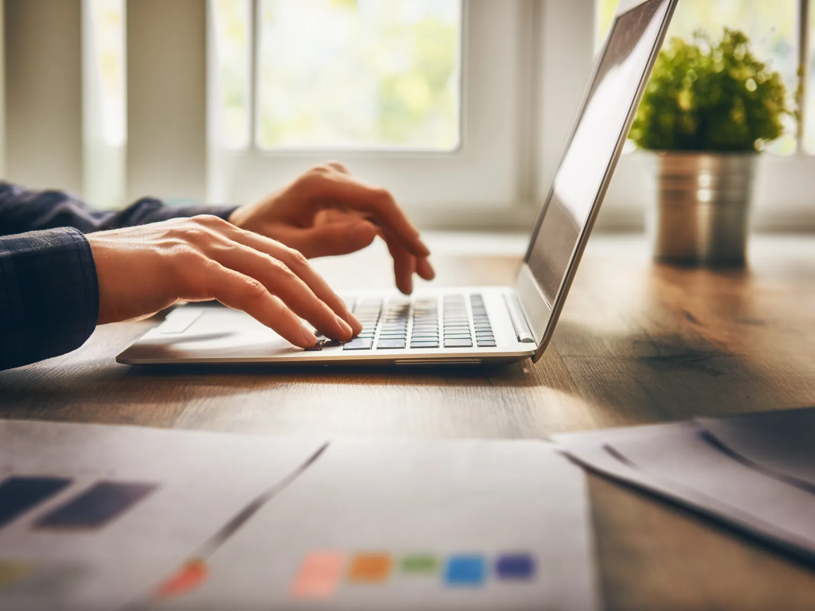 Person typing on a laptop keyboard with papers and a small plant on a wooden desk near a window.