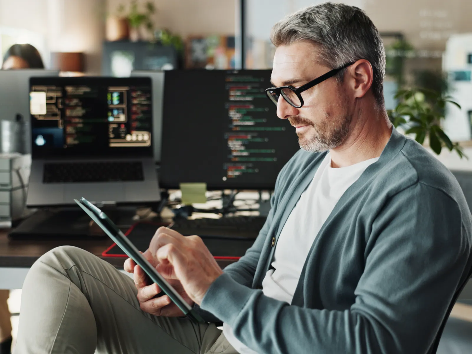 Man in glasses using tablet in modern office with multiple computer screens showing code in the background