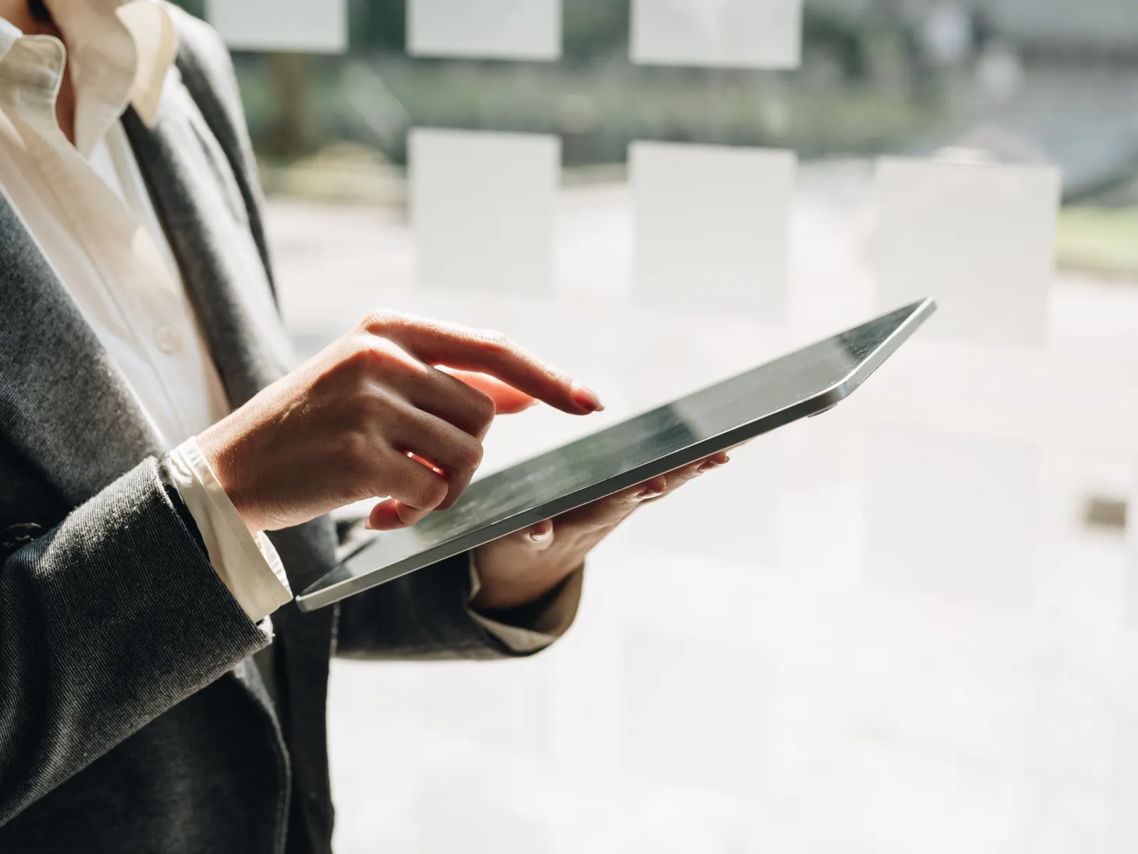 Businessperson in suit using a digital tablet with a blurred window background, focusing on touch interaction.
