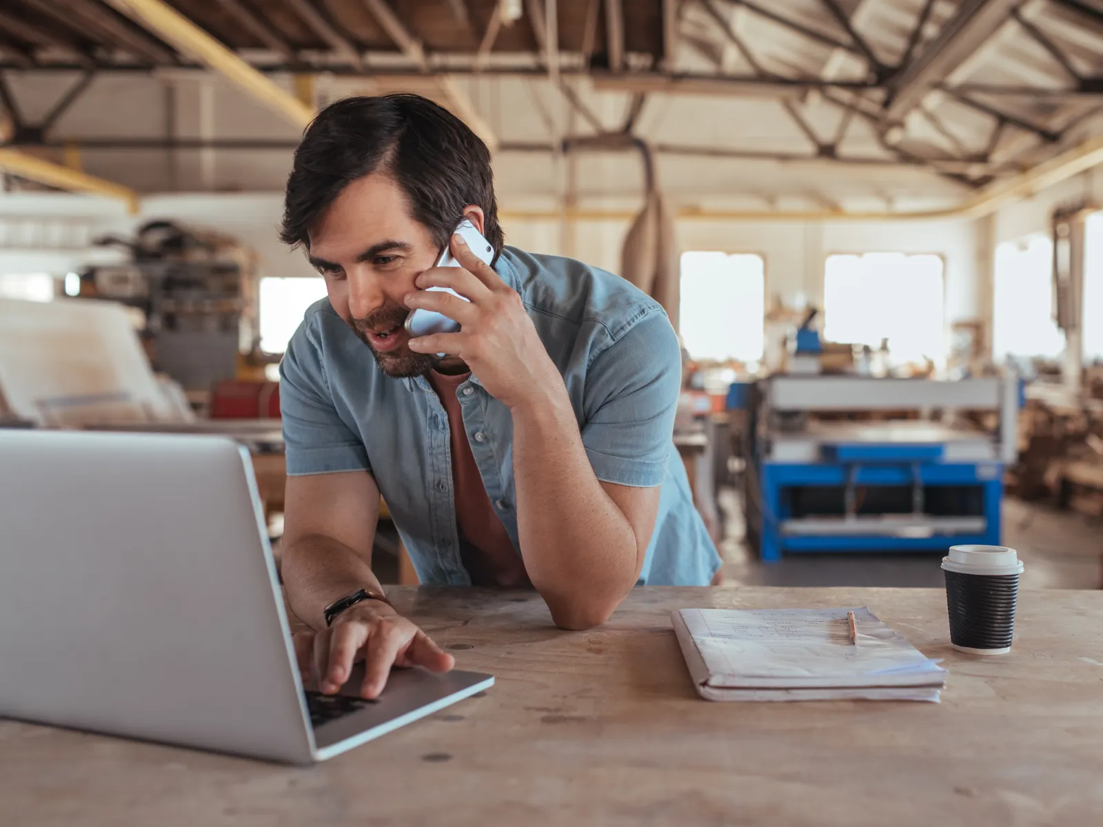 Man in a workshop using a laptop and talking on a phone with a coffee cup and plans on the table.