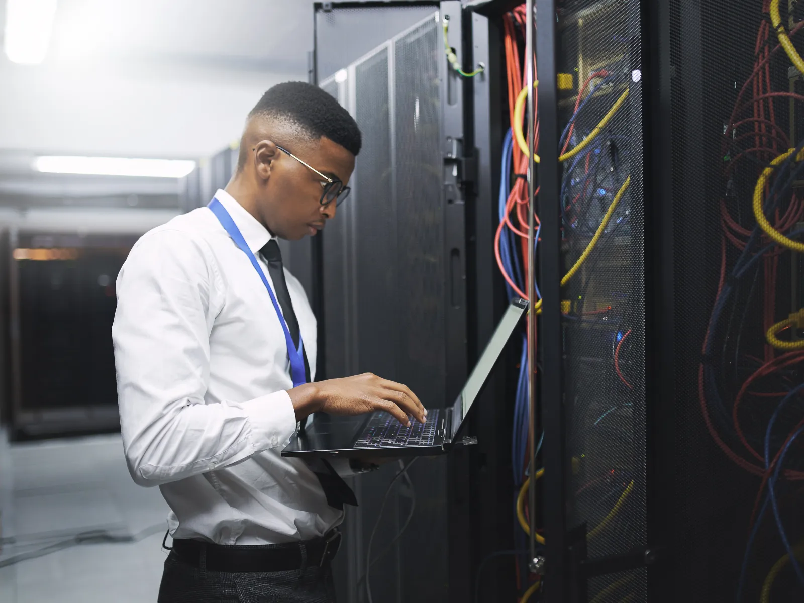 IT technician in formal attire working on a laptop in a data center server room with network cables.