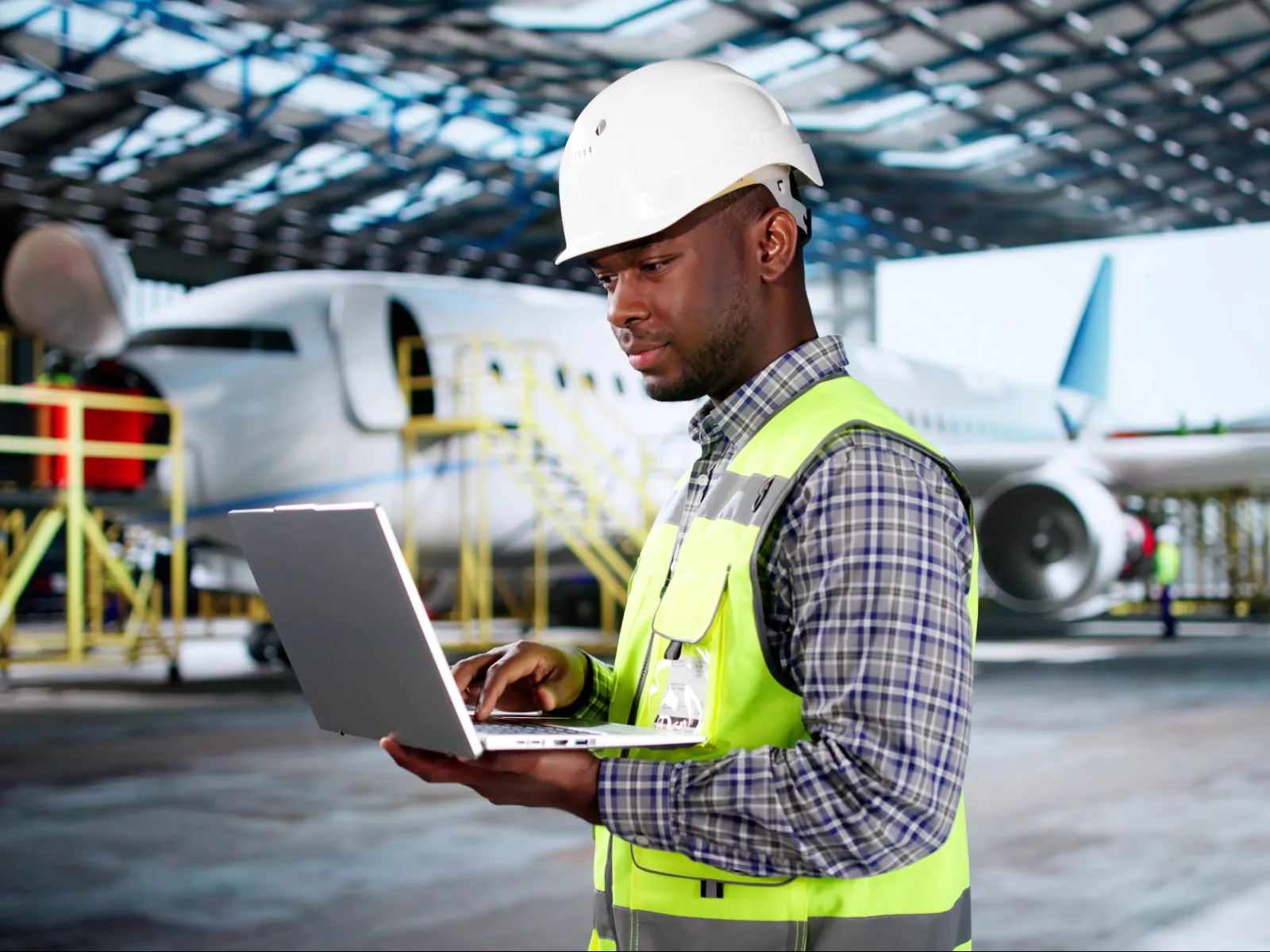 Engineer in safety vest and helmet using laptop inside airplane hangar with jet in background under large roof.
