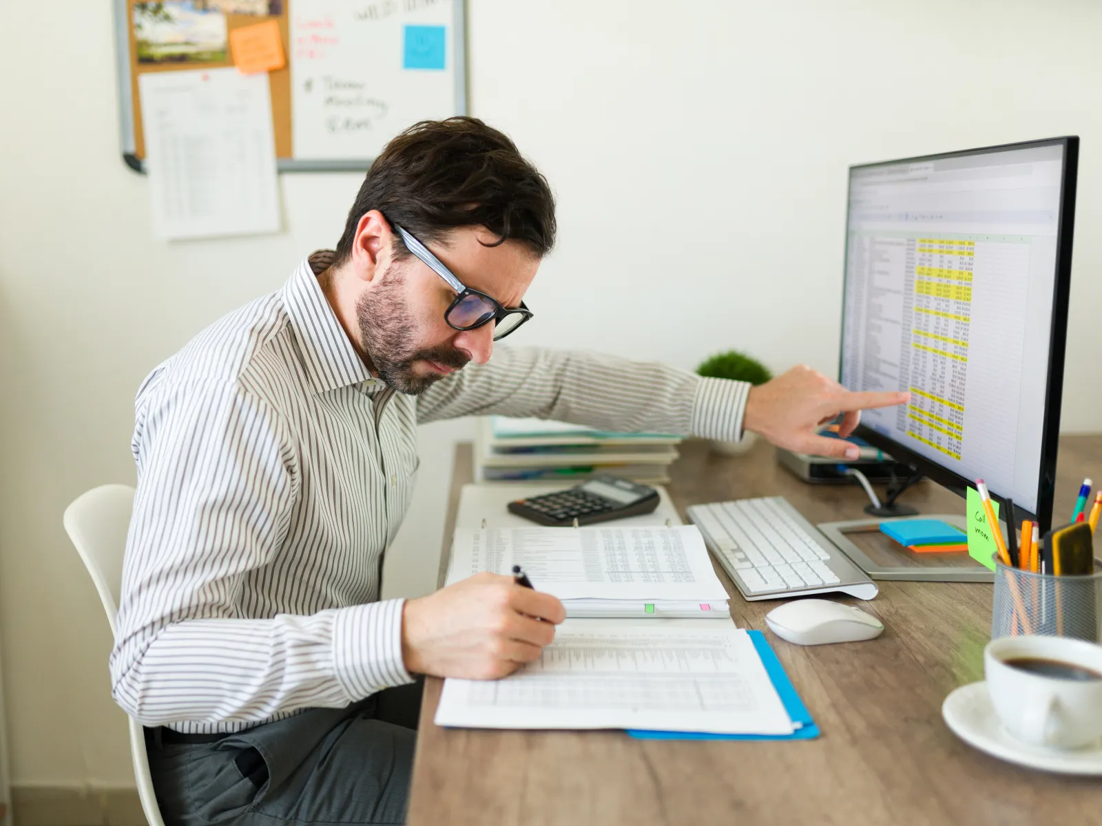 Man in glasses working at desk reviewing financial documents and spreadsheet on computer screen