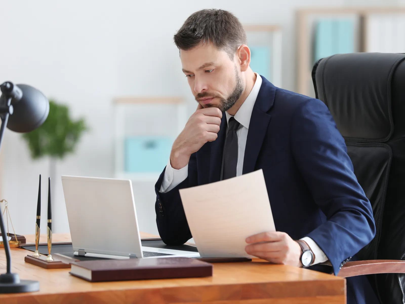 Businessman in a suit thoughtfully reviewing documents at a desk with laptop and office supplies.