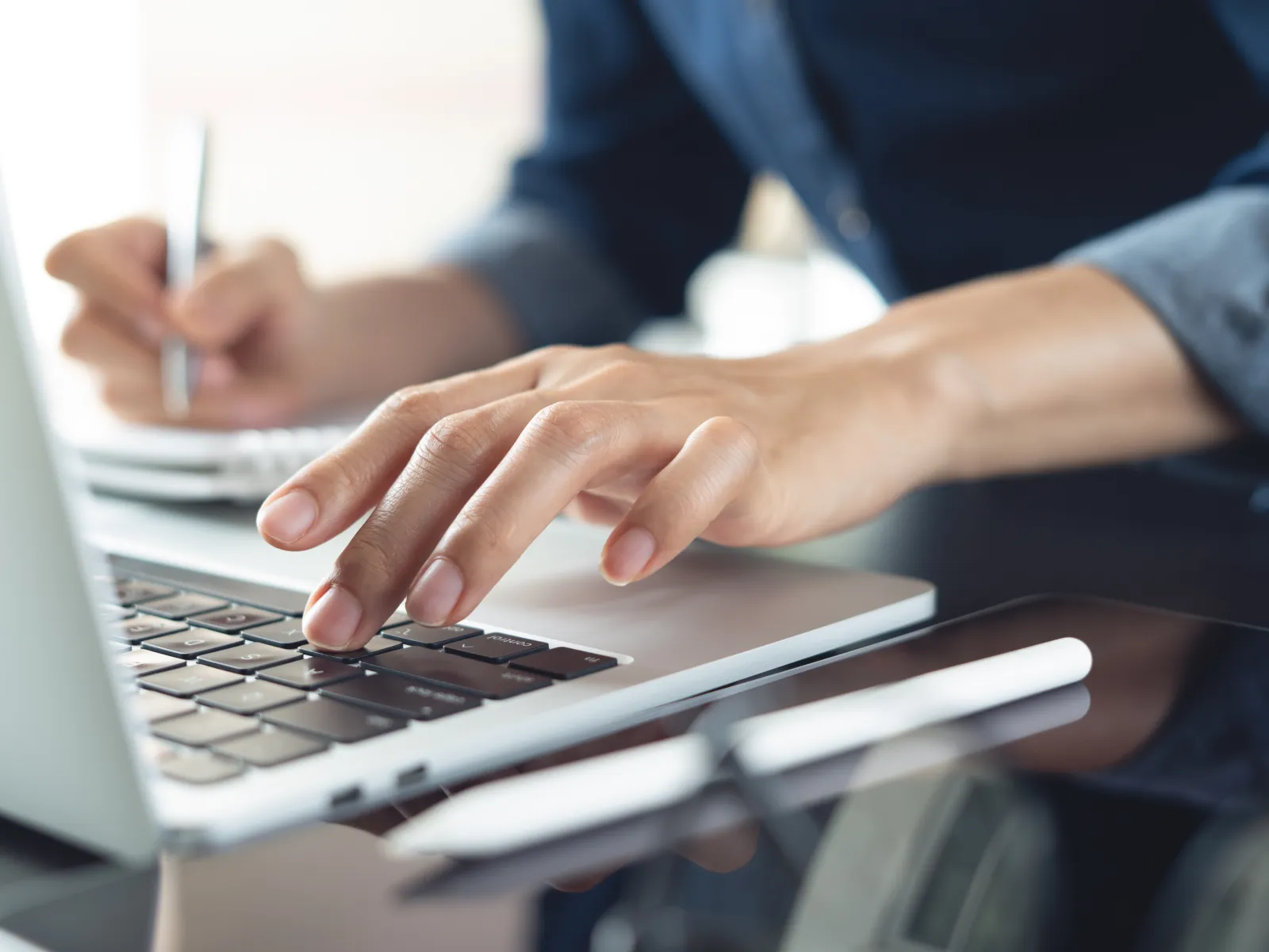 Close-up of a person typing on a laptop keyboard while taking notes with a pen on paper.