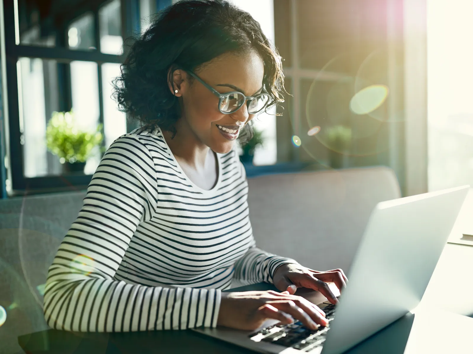 Smiling woman wearing glasses working on a laptop in a bright room with sunlight and plants.