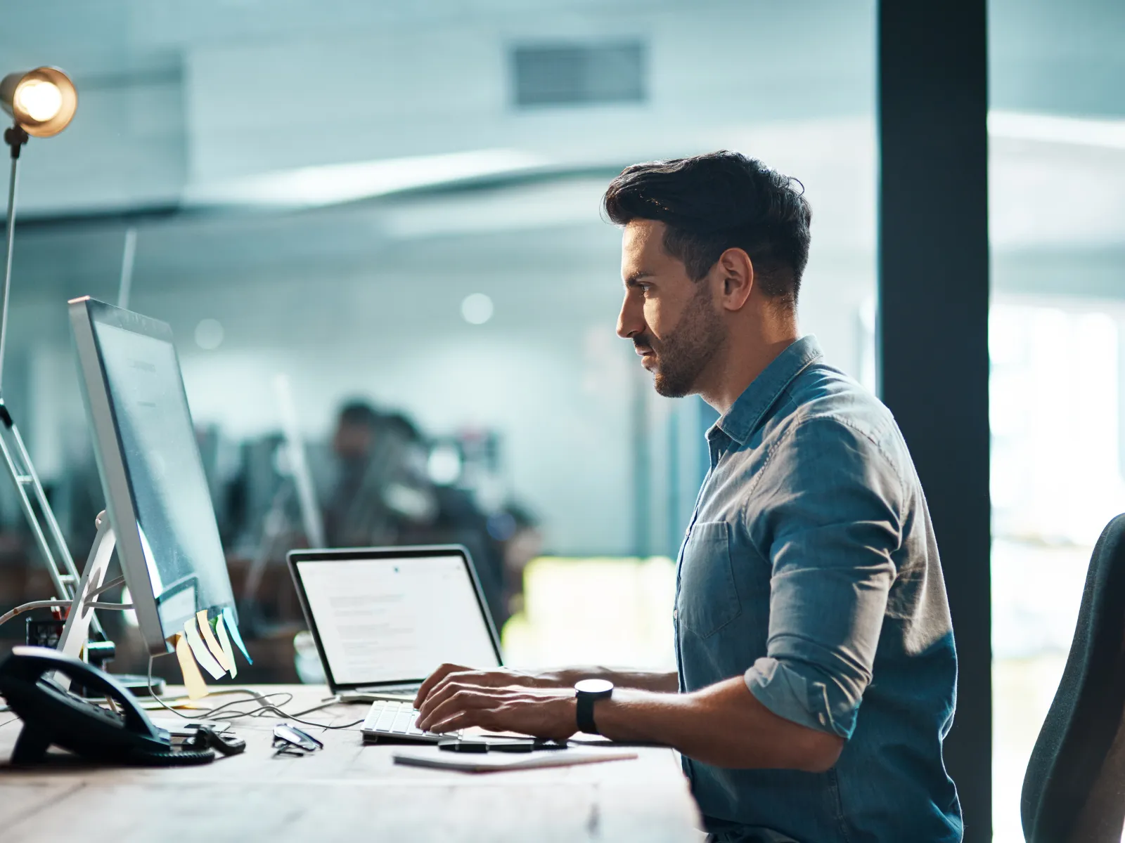 Man in blue shirt working on desktop computer in modern office with laptop and phone on desk