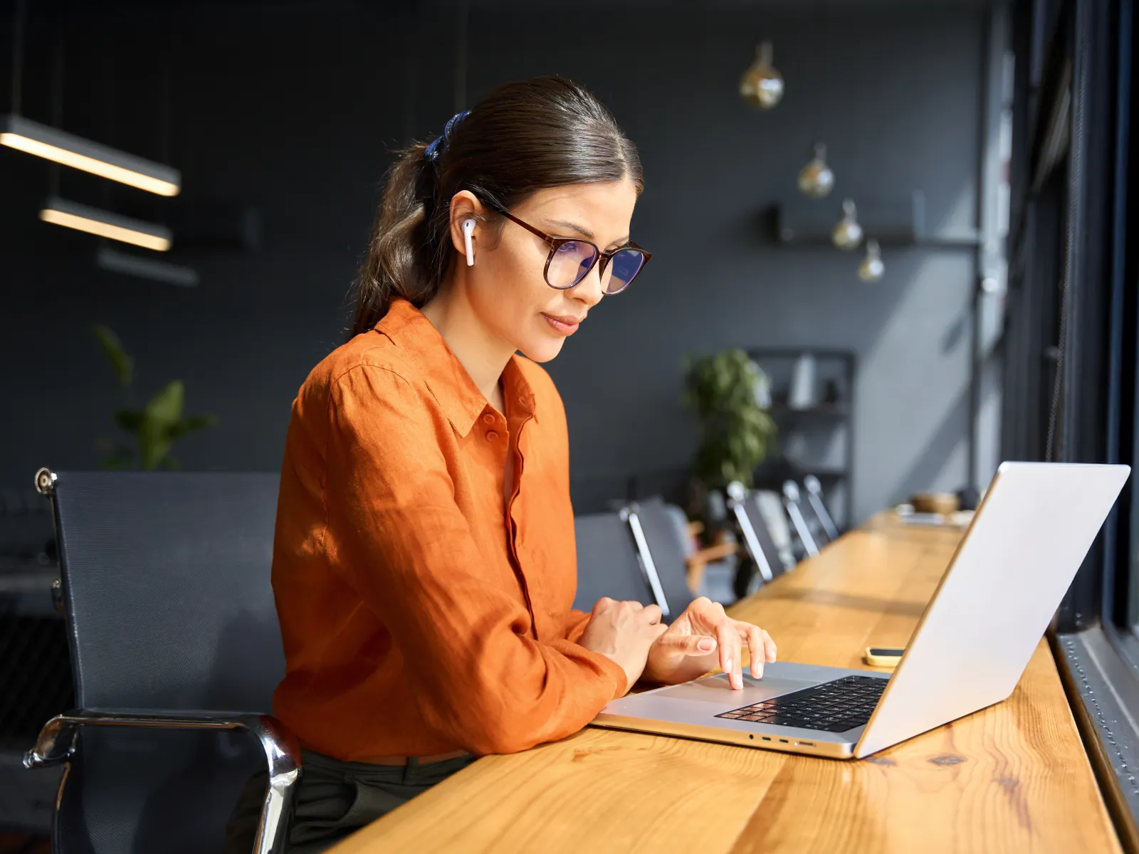 Young woman in orange shirt working on laptop in a modern cafe with natural light from large windows.