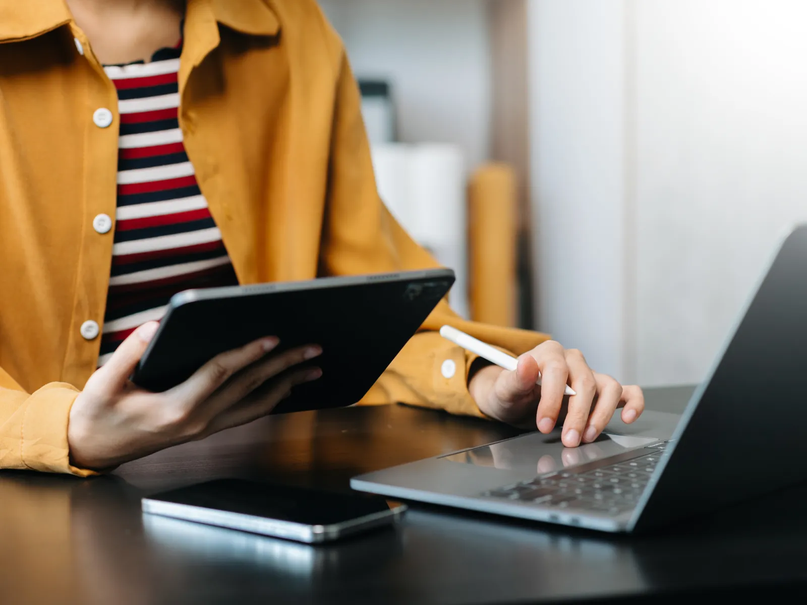Person using tablet and laptop with smartphone on desk, multitasking in modern workspace.
