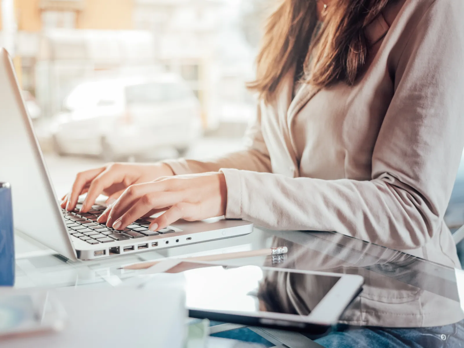 Close-up of a woman typing on a laptop at a glass desk with a tablet and coffee mug nearby