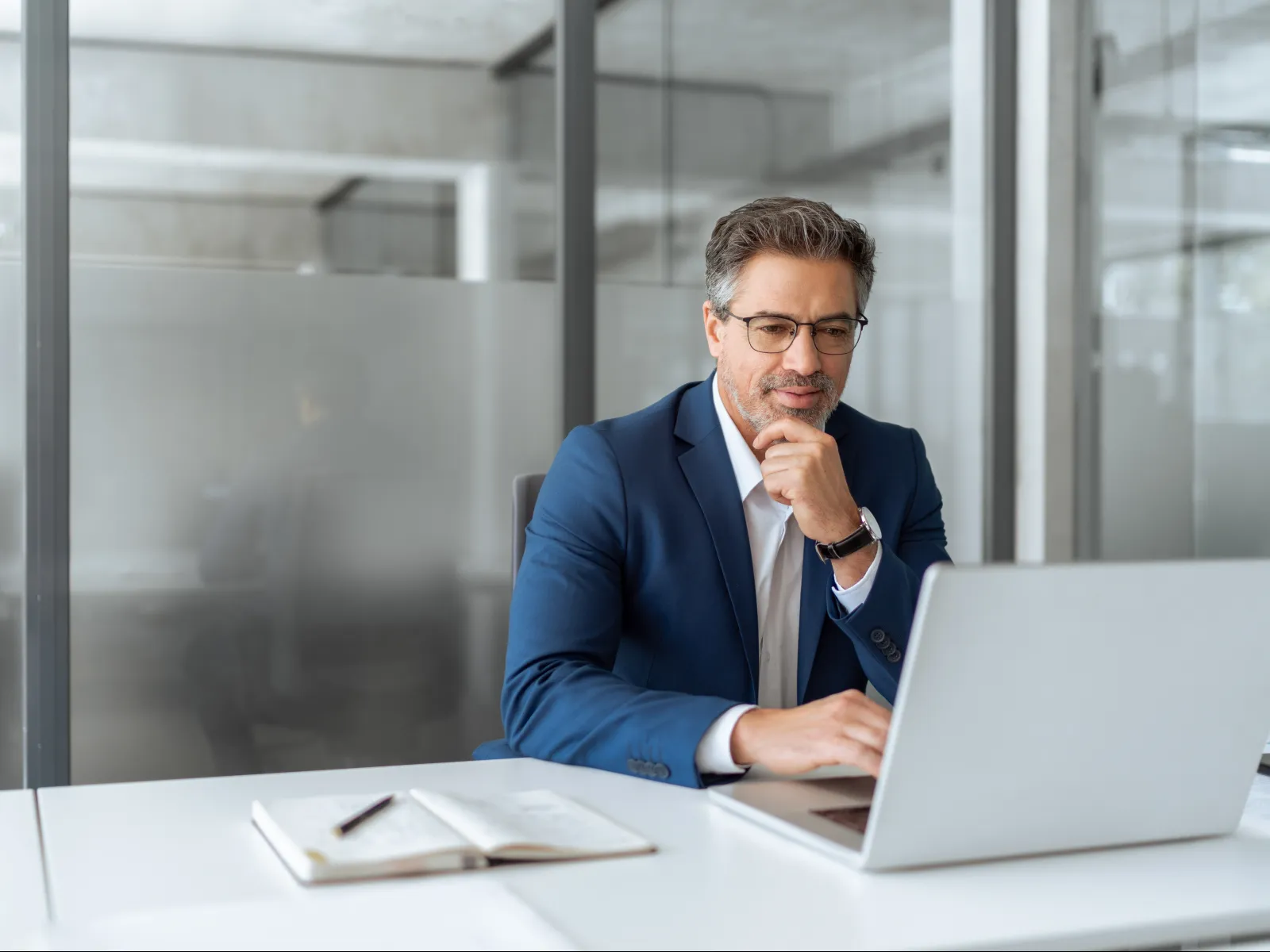 Professional businessman in blue suit working thoughtfully on laptop in modern office with notebook nearby