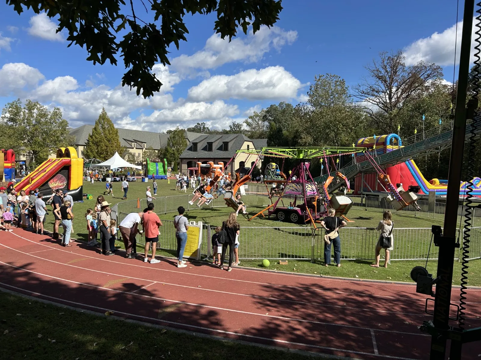 Families enjoy sunny day at outdoor carnival with inflatable slides and swing rides on green field beside running track.