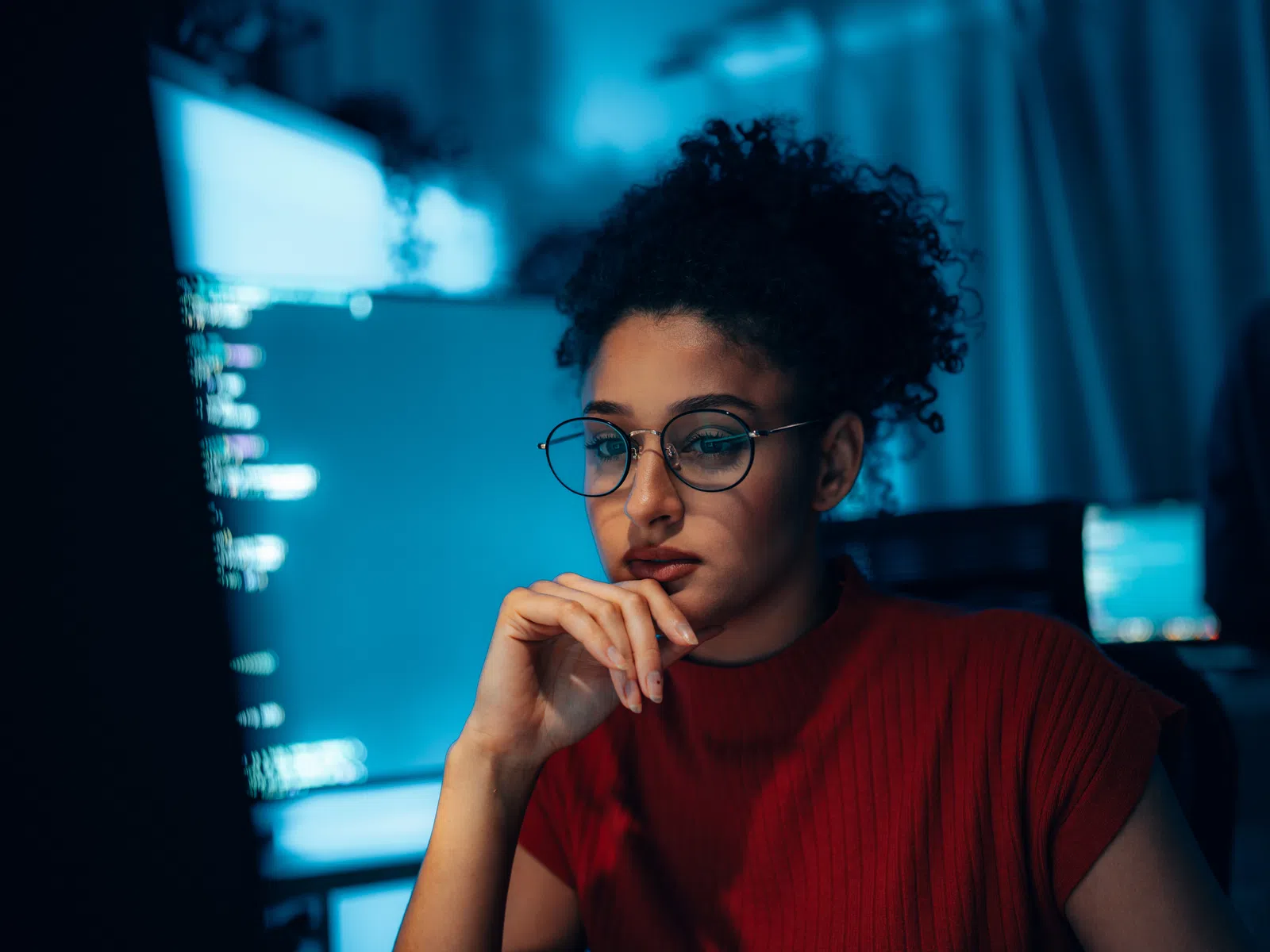 Young woman in glasses and red sweater thoughtfully coding in front of a computer screen at night.