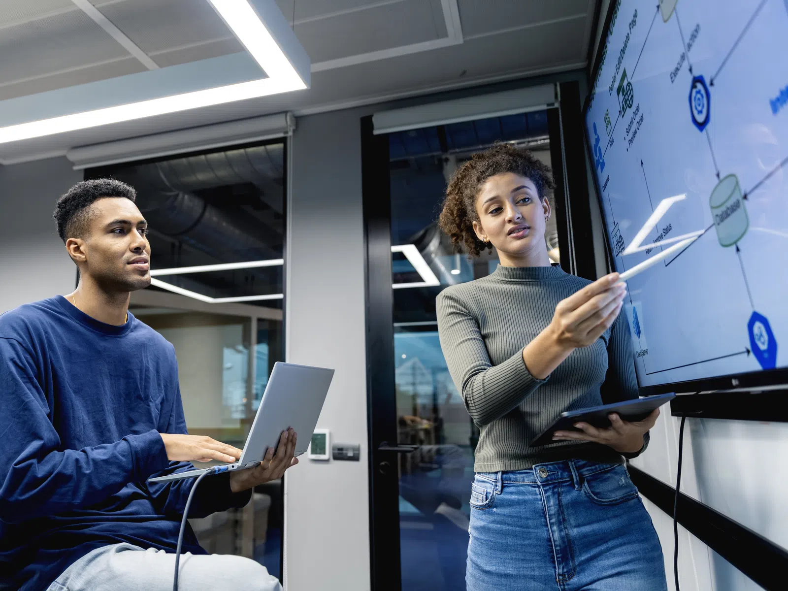 Two colleagues discussing data on a large screen in a modern office, one holding a laptop and the other pointing.