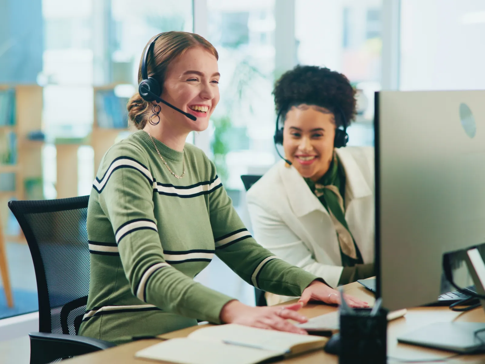 Two women wearing headsets working together at a computer in a bright modern office environment