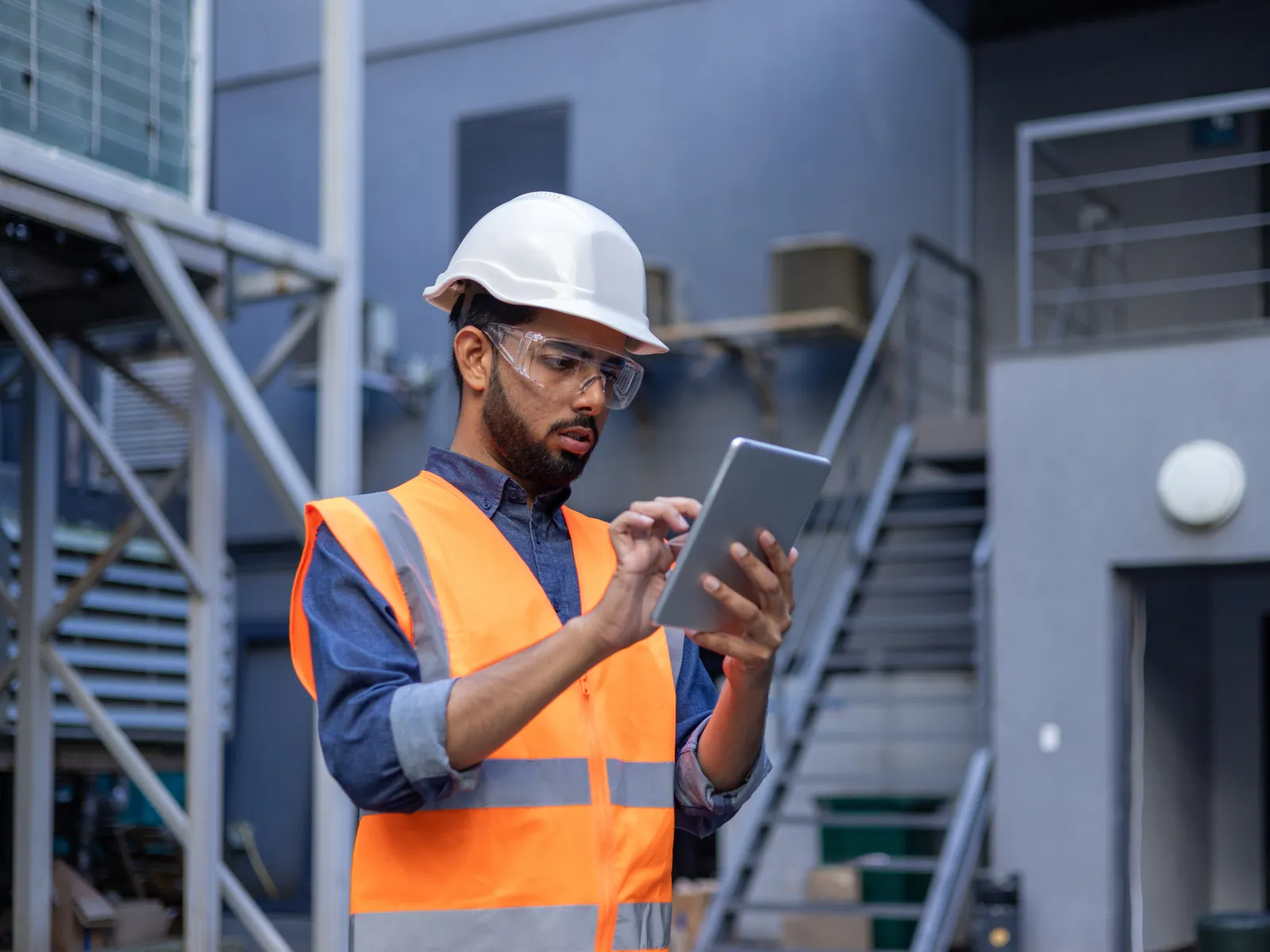 Frustrated worker in safety gear, holding helmet, looking at laptop in a warehouse setting.