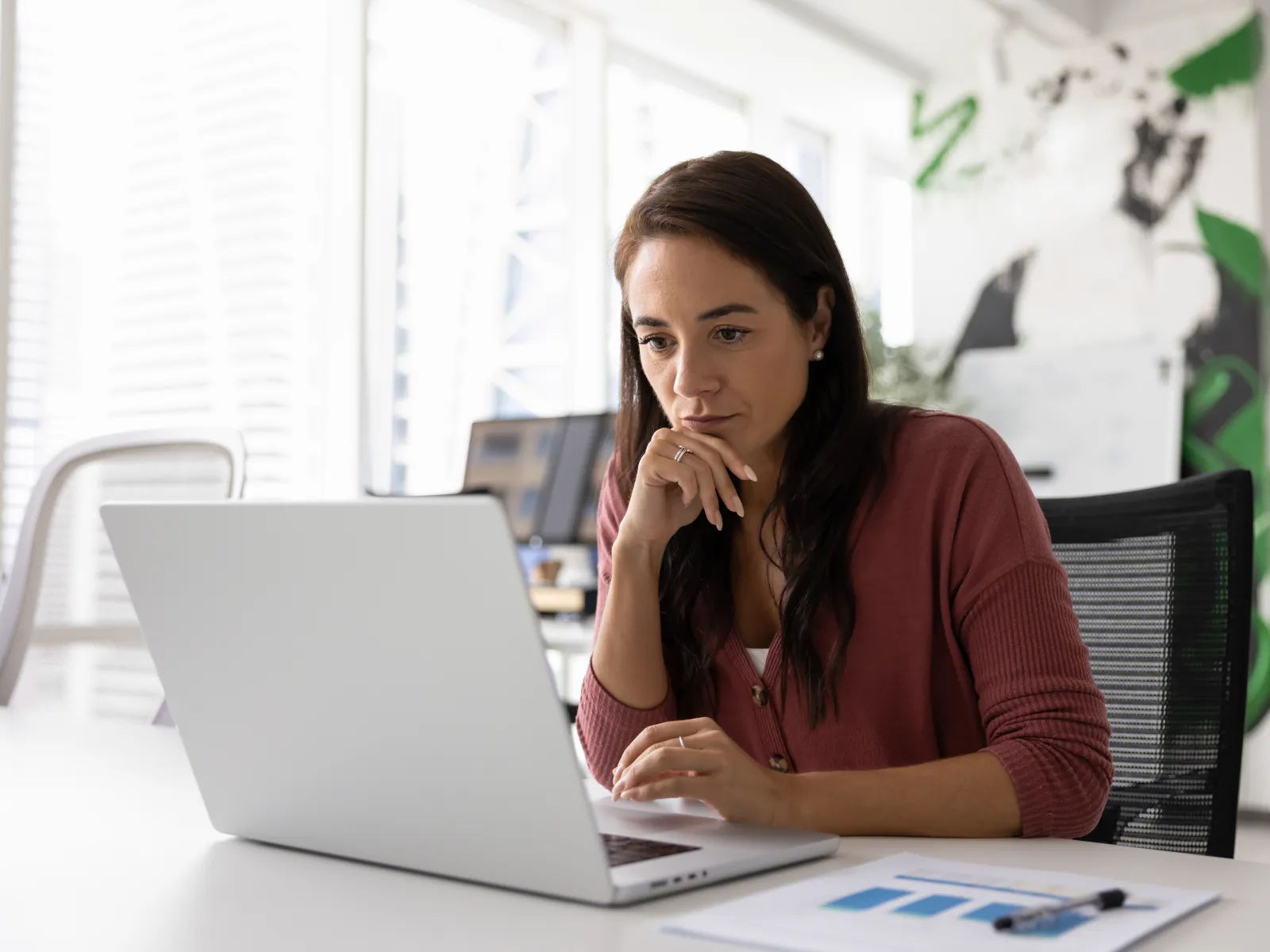 Young woman in casual shirt focused on working with laptop in bright modern office space