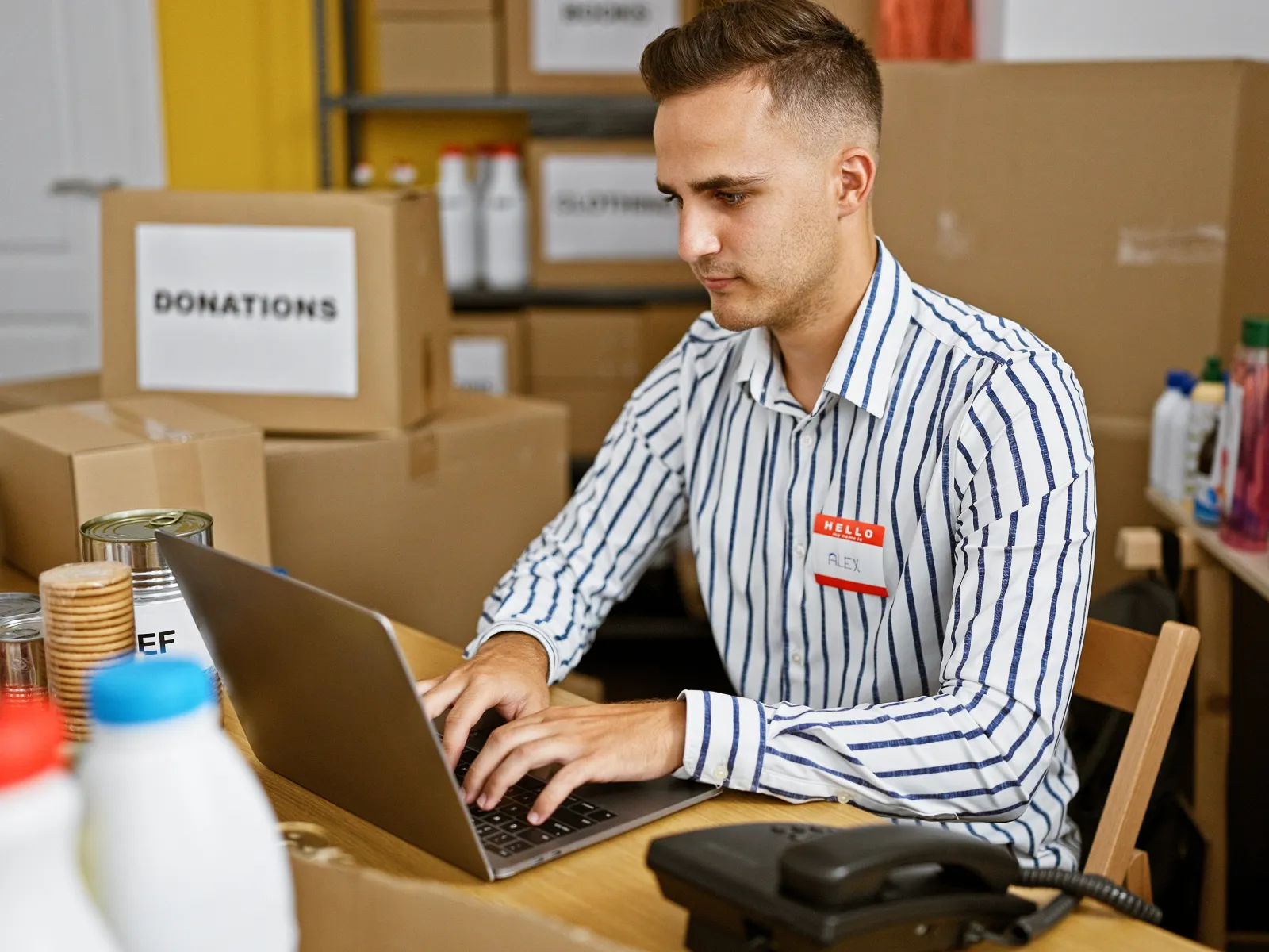 Volunteer with name tag working on laptop surrounded by donation boxes and food supplies in a charity center.