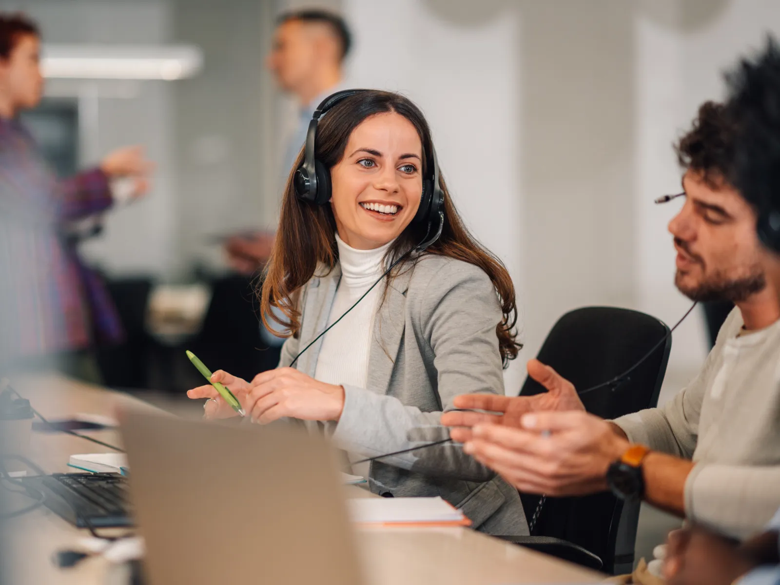 Smiling woman with headset talking to colleagues in an office call center environment.