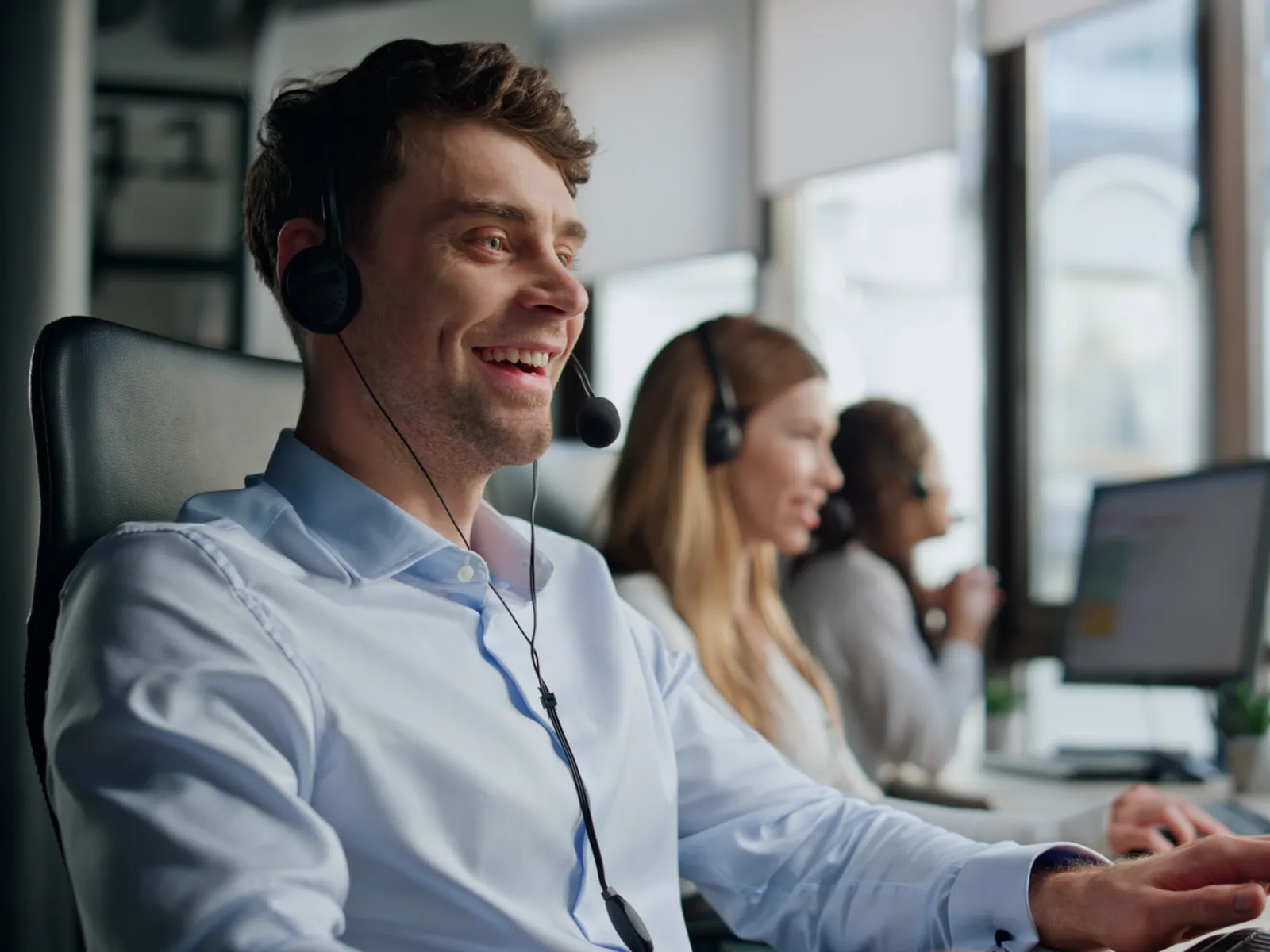 Smiling male customer service agent wearing headset working on computer in bright modern office with colleagues.