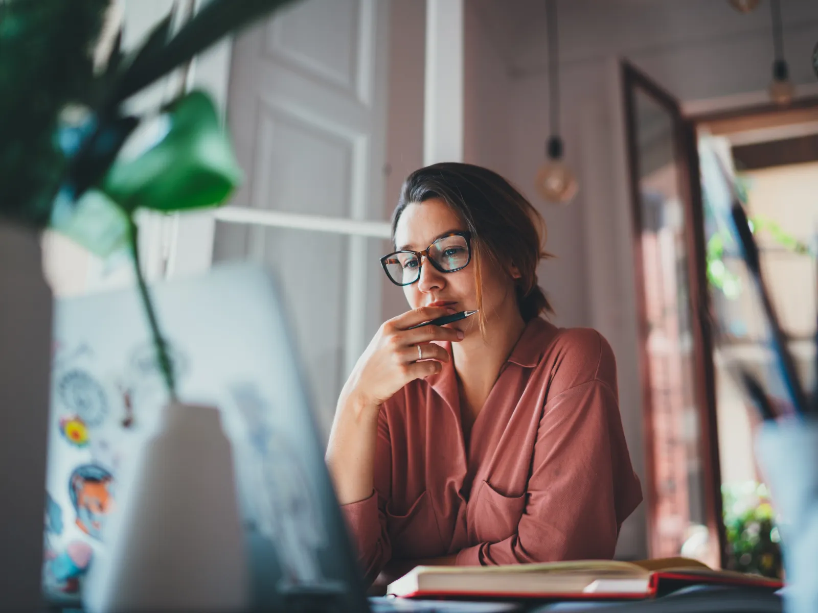 Woman wearing glasses and a pink shirt thoughtfully working on a laptop in a cozy, sunlit room.