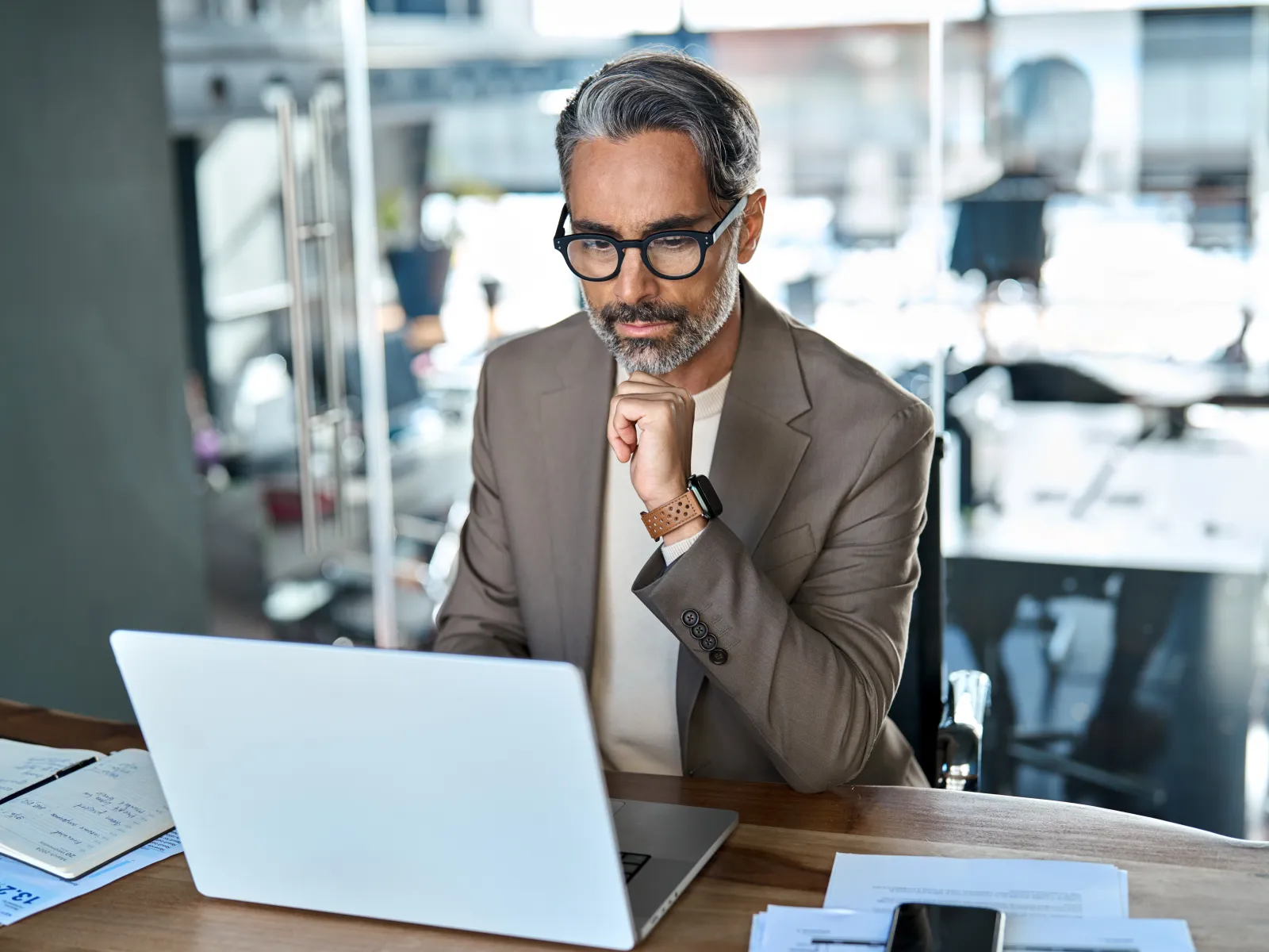 Middle-aged man with gray hair and glasses working focused on a laptop at a modern office desk.