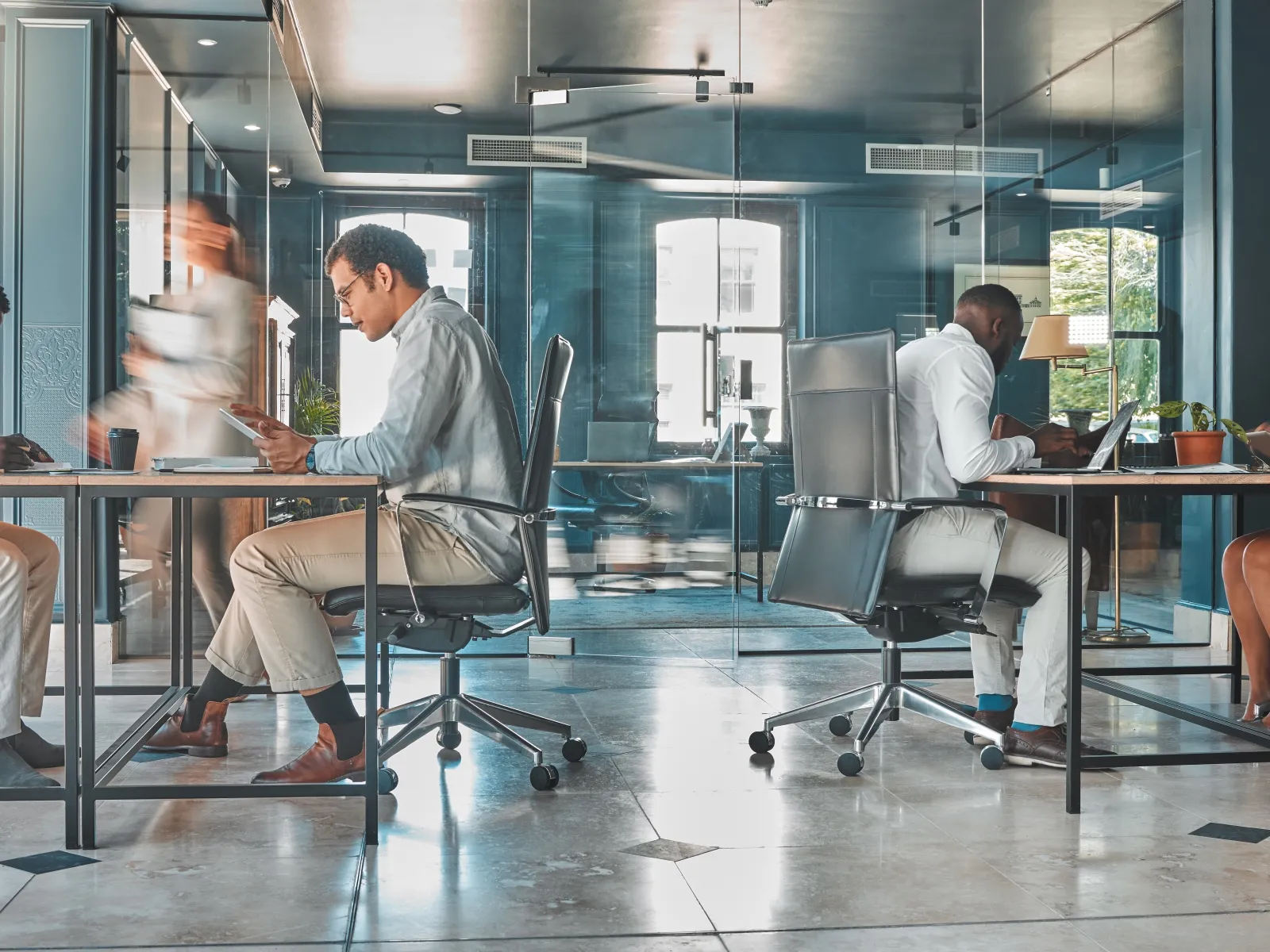 Diverse professionals working on laptops and phones in a modern glass-walled office space with natural light.