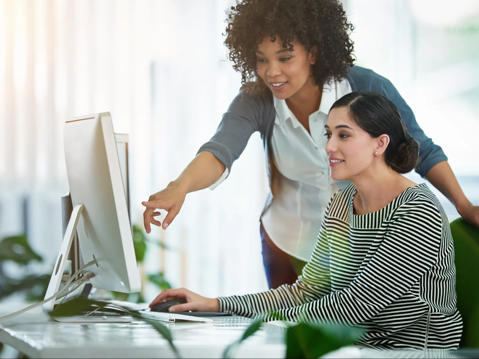 Two women collaborating at a computer in a bright modern office with natural light and plants.