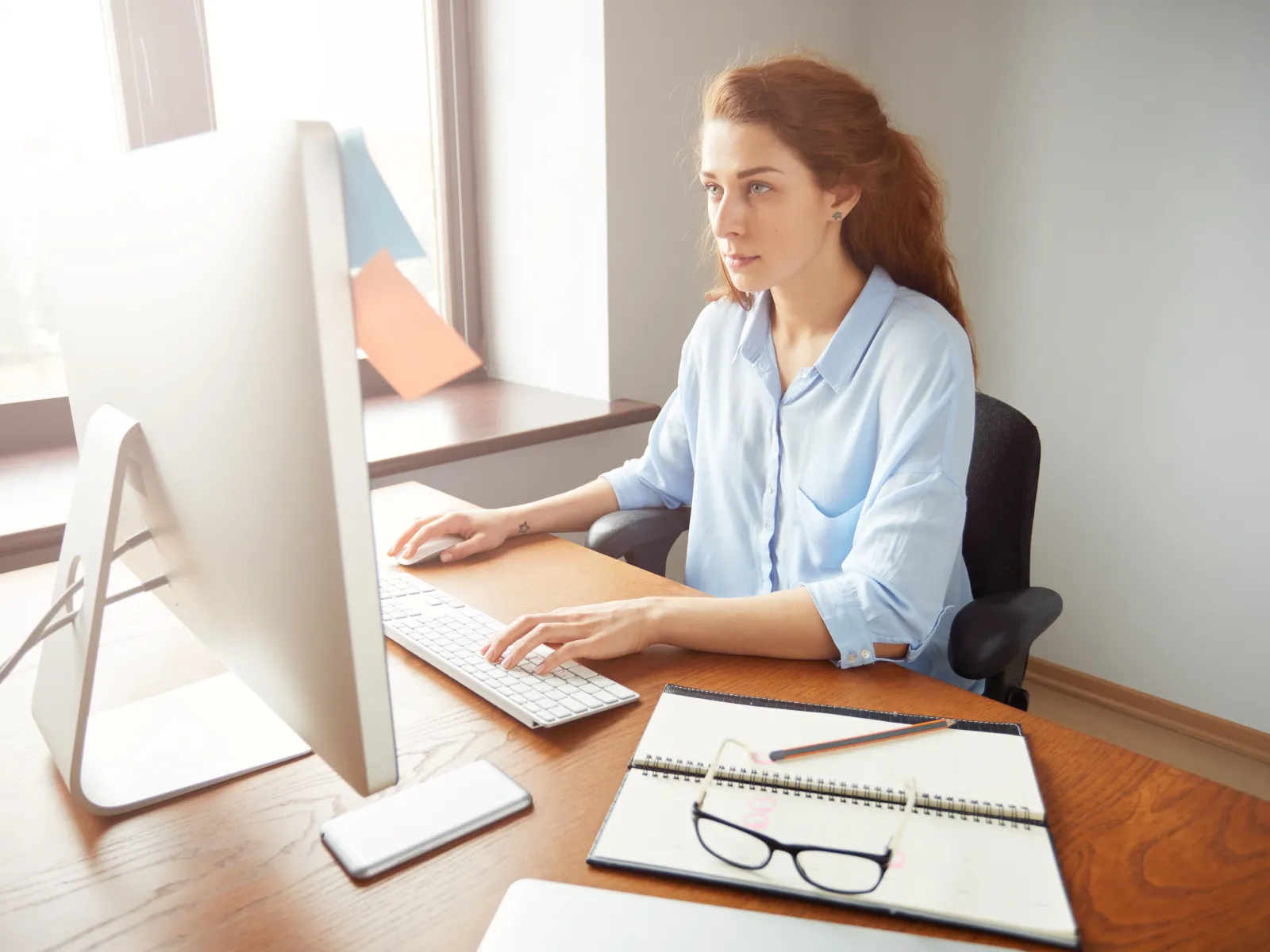 Young woman focused on computer work at wooden desk with notebook, glasses, and smartphone near window