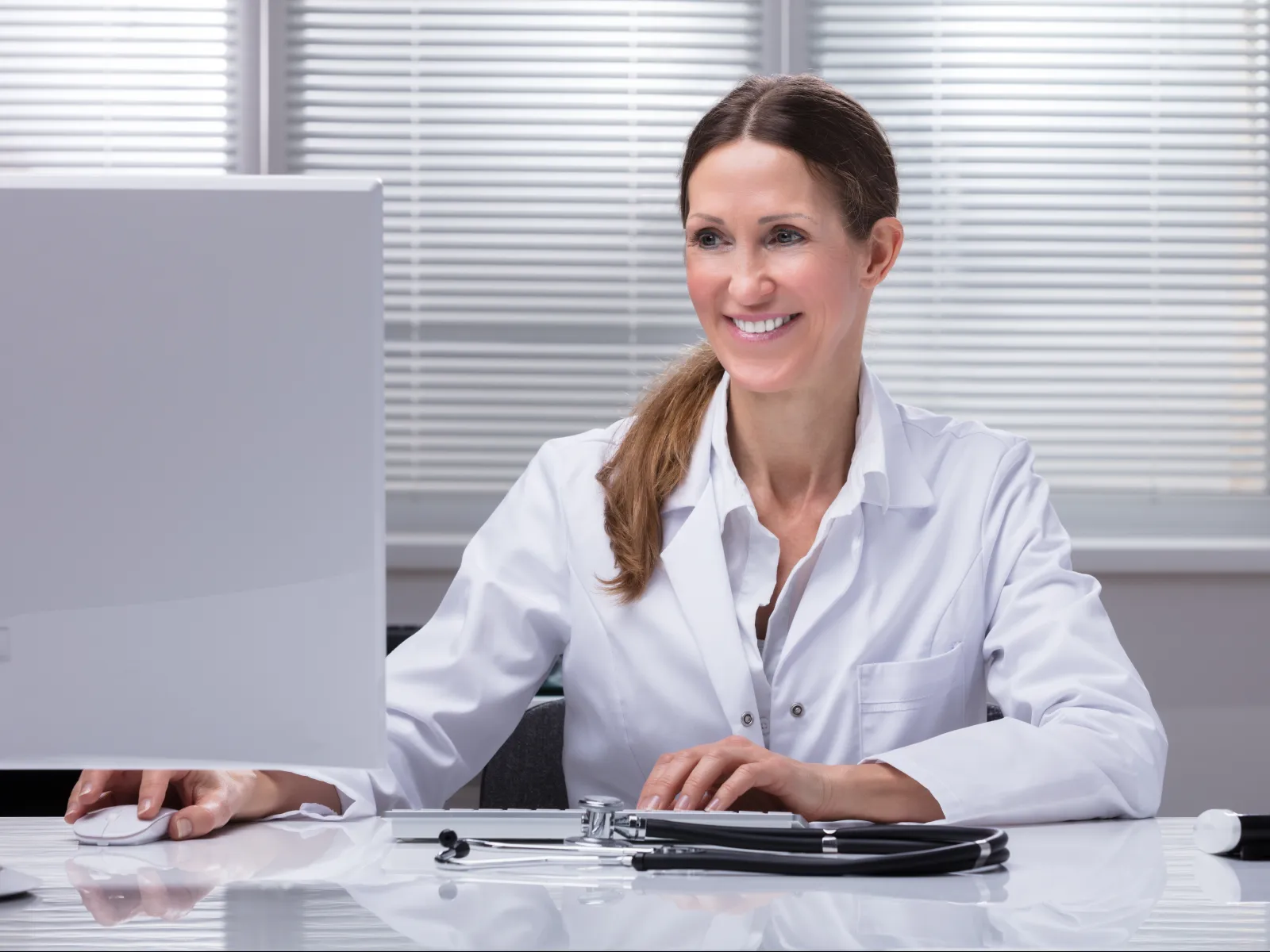 Smiling female doctor in white coat using computer in modern medical office with stethoscope on desk