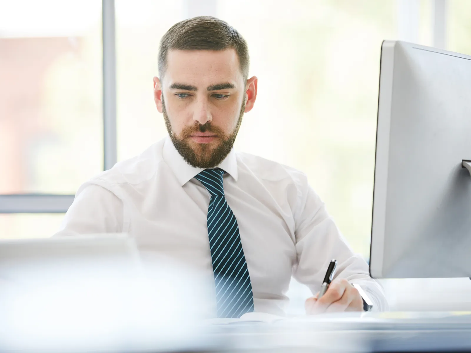 Businessman in white shirt and striped tie working at desk with digital tablet and computer in bright office