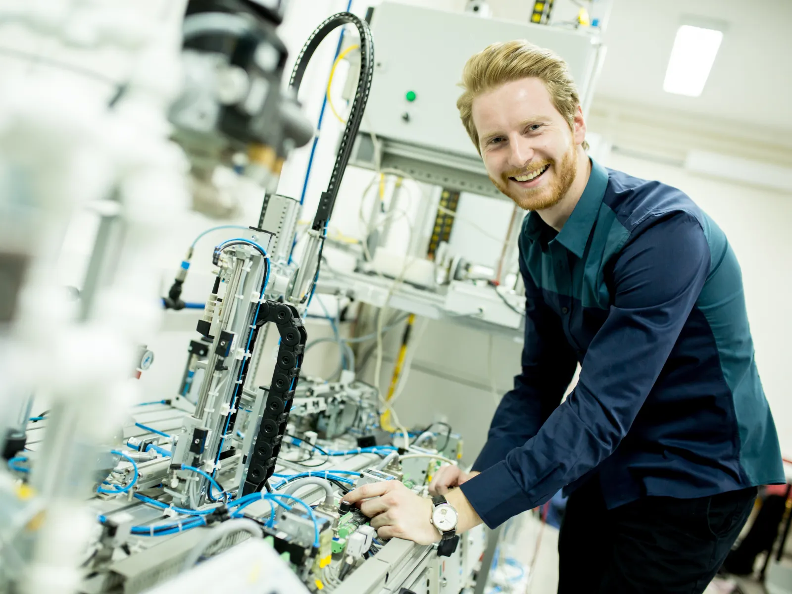 Smiling technician adjusting complex industrial automation equipment with wires and pneumatic components.