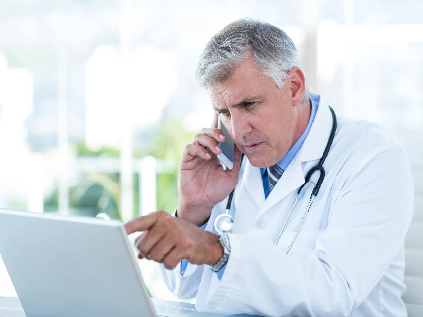 Focused male doctor in white coat using laptop and talking on smartphone in bright modern office.