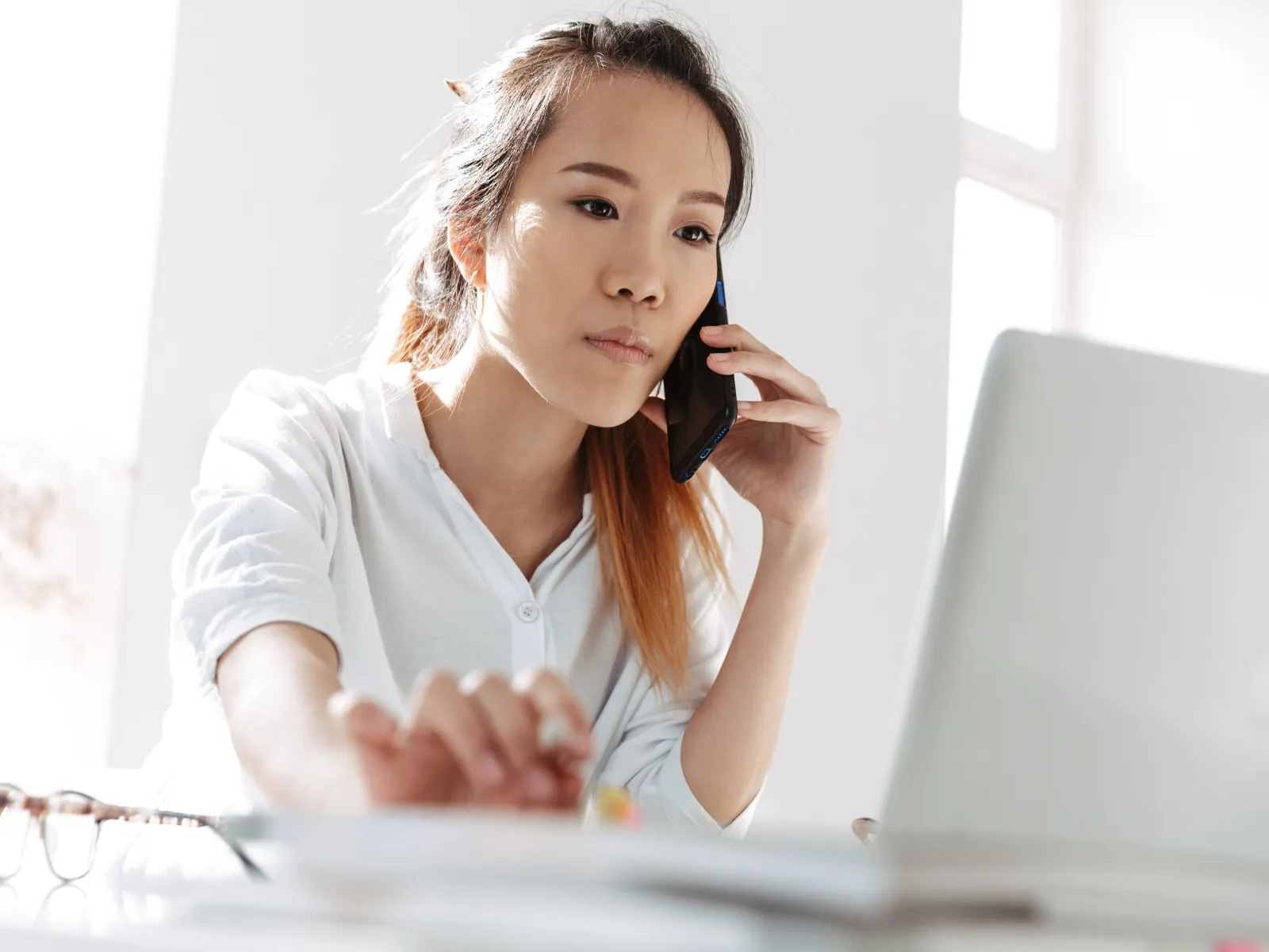 Young woman in white shirt talking on phone while using laptop at bright workspace