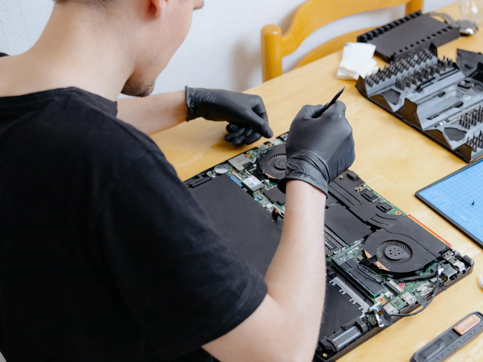 Technician wearing black gloves repairing laptop motherboard on wooden table with tools nearby.