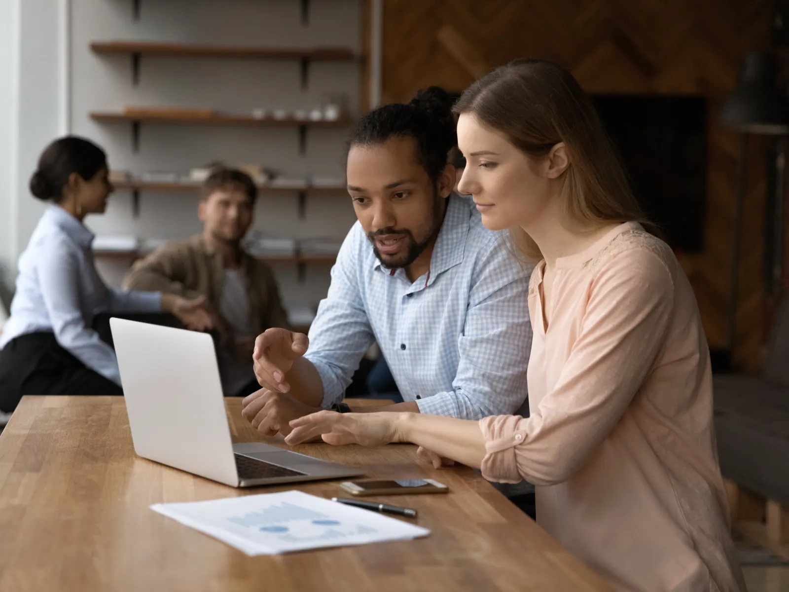 Two coworkers collaborating over a laptop at a wooden table in a modern office setting.