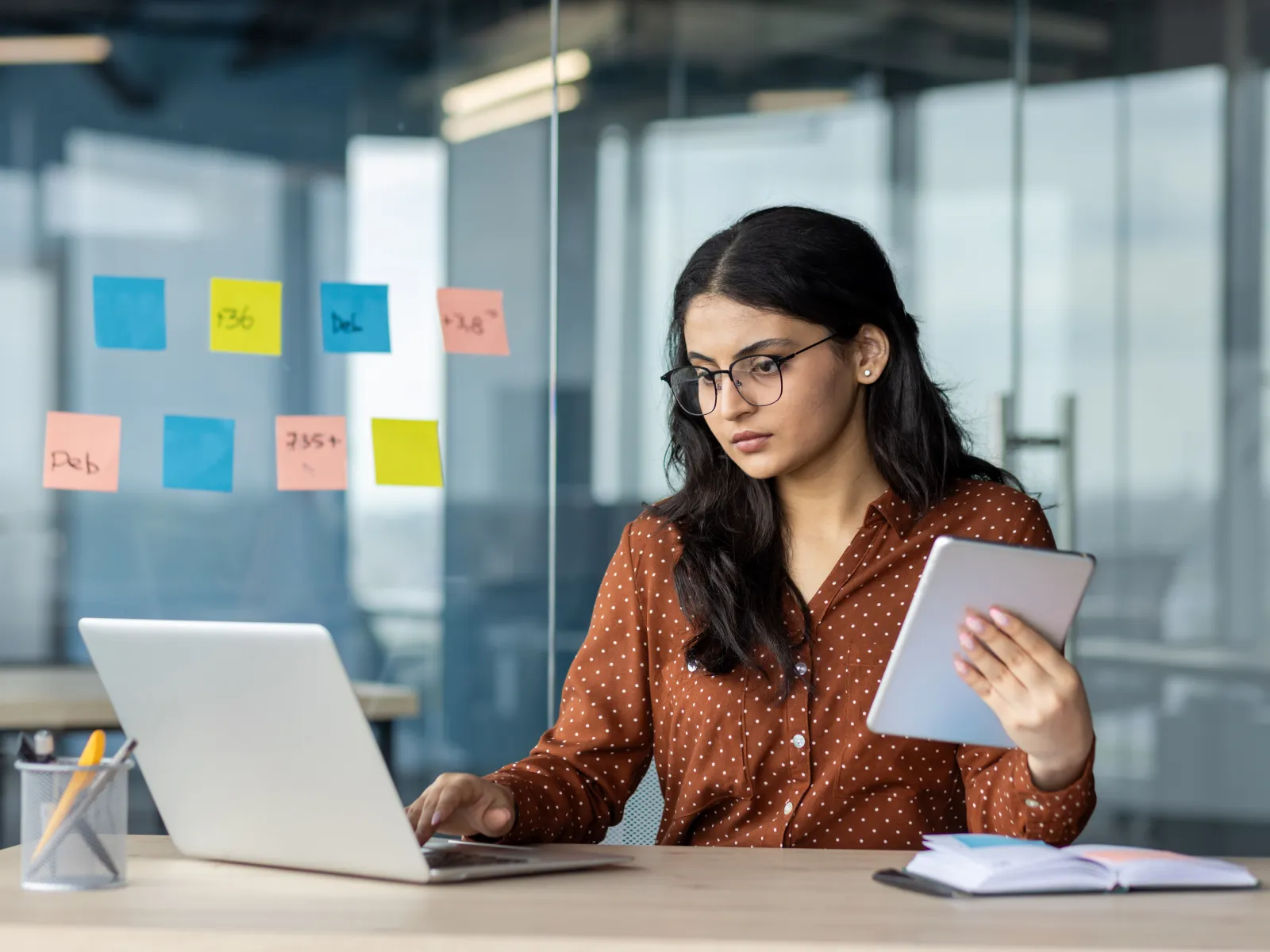 Young woman working on laptop and tablet in modern office with colorful sticky notes on glass wall.