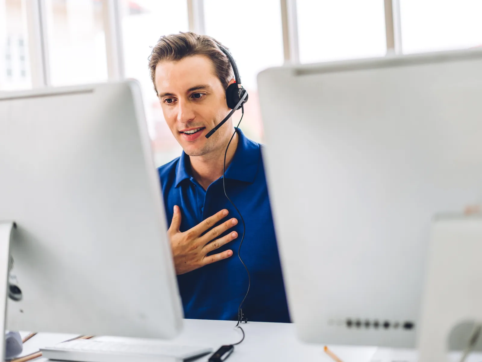 Customer service representative with headset speaking and gesturing in front of two computer monitors.