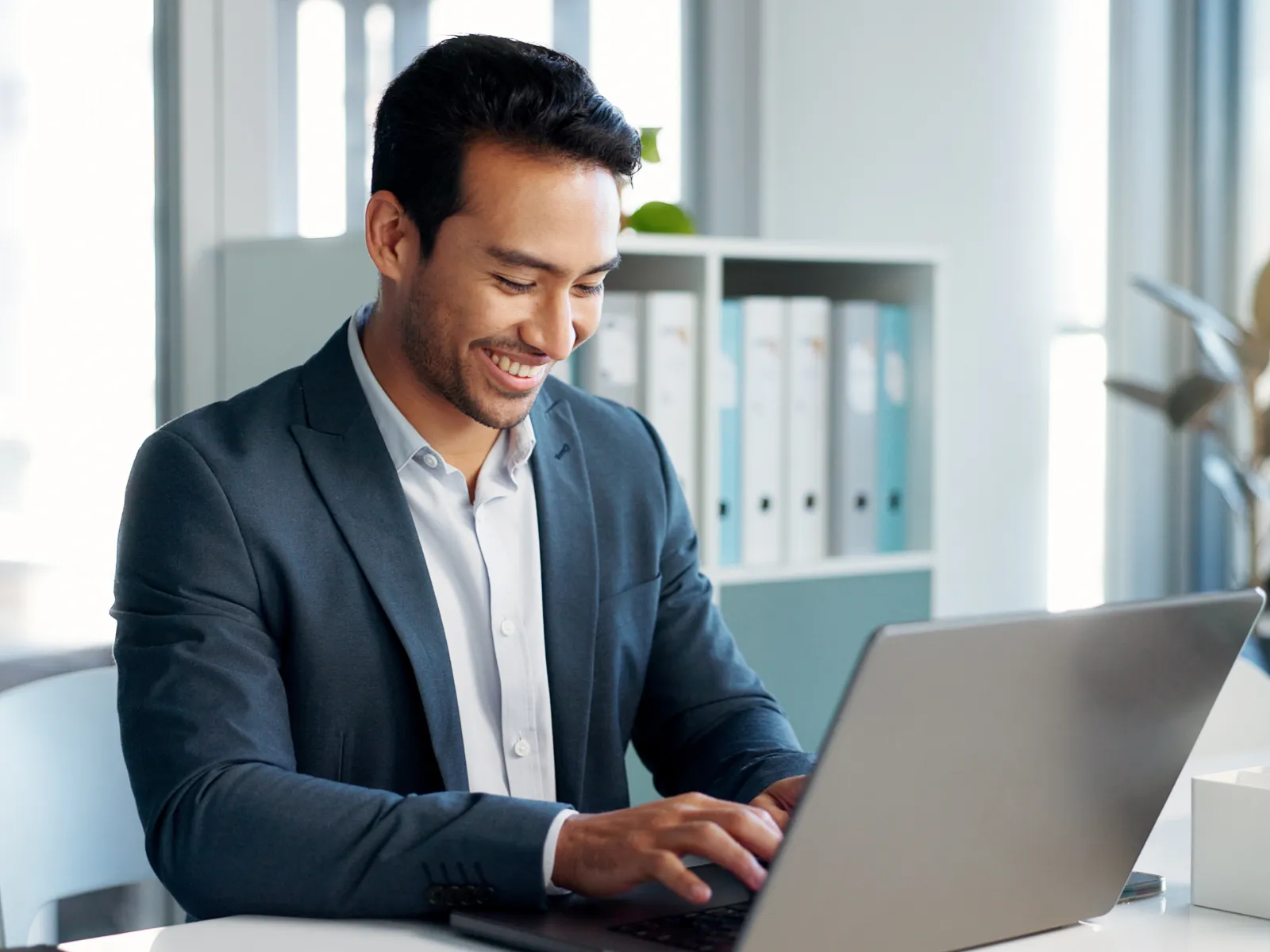 Smiling businessman in suit working on laptop at modern office desk with bright background light