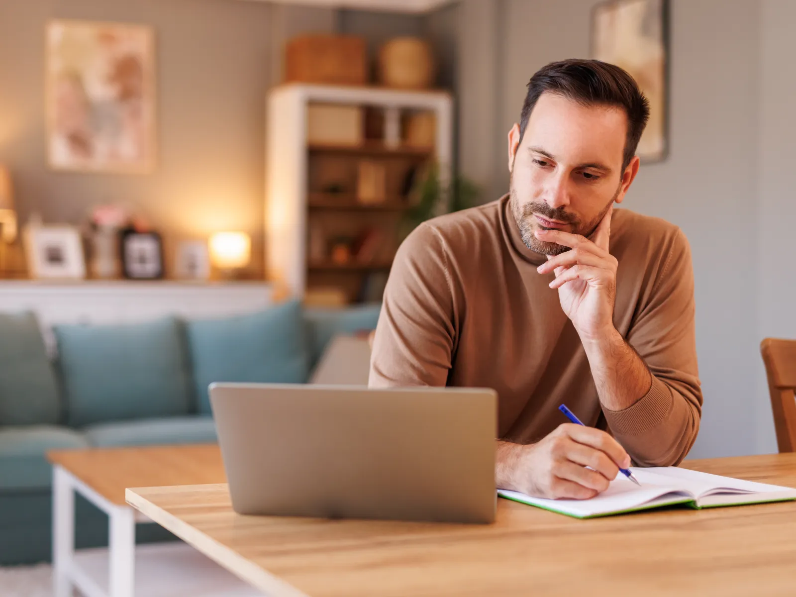 Man thoughtfully working on laptop and writing notes at wooden table in cozy living room