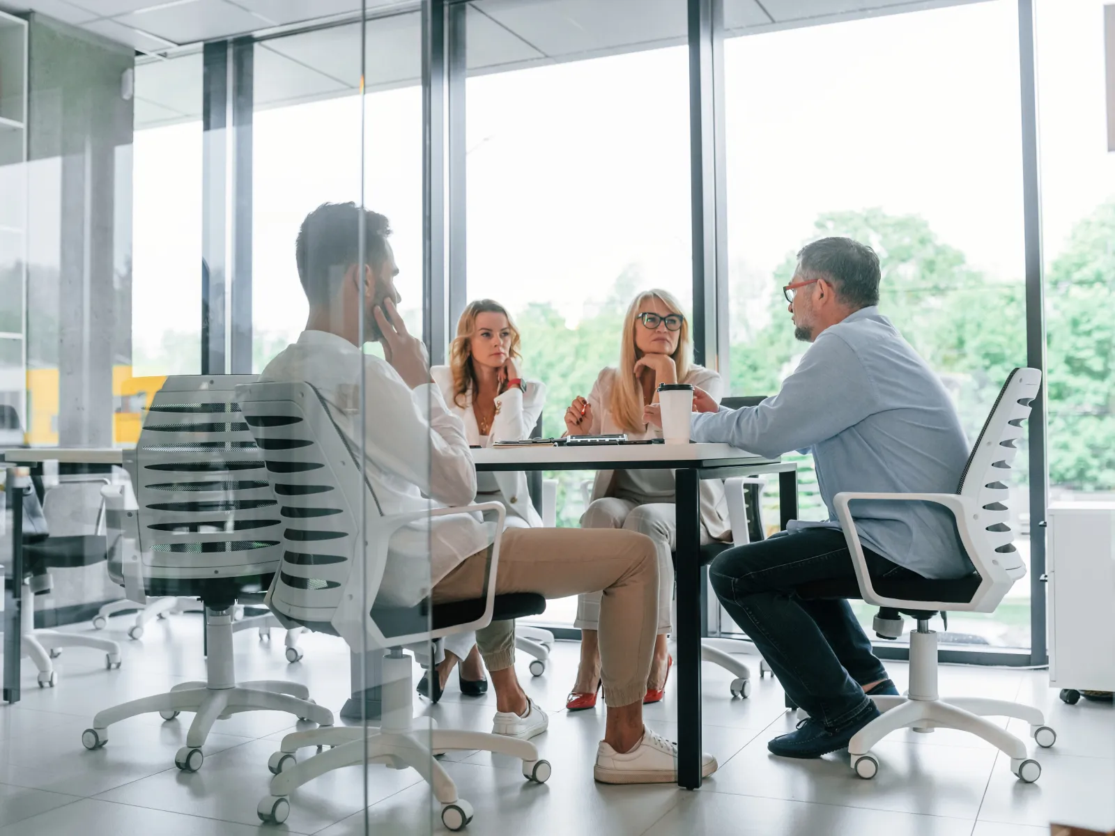 Four professionals engaged in a discussion around a table in a bright, modern office with glass walls.