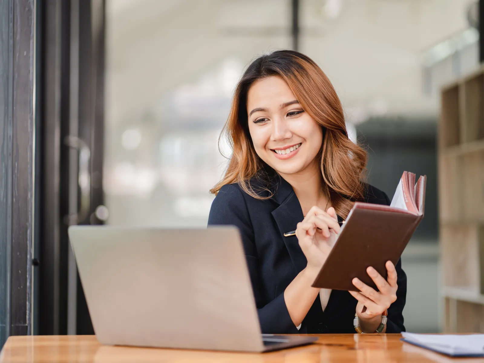 Smiling woman in business attire taking notes while working on a laptop at a wooden desk in modern office.
