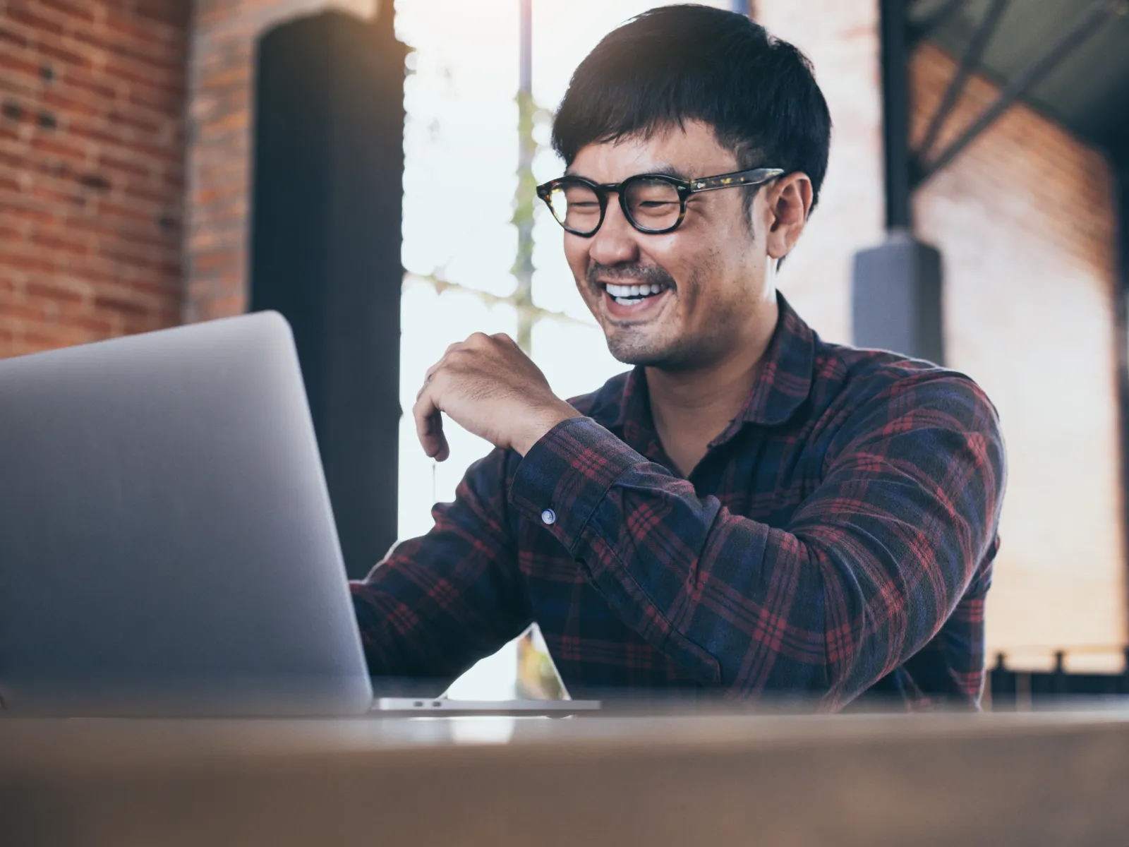Smiling man wearing glasses works on laptop in bright modern office with brick walls and natural light