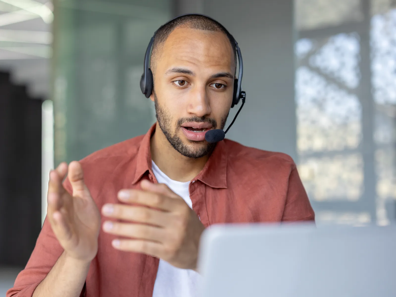 Man wearing headset and orange shirt talking and gesturing during a video call on laptop in modern office.